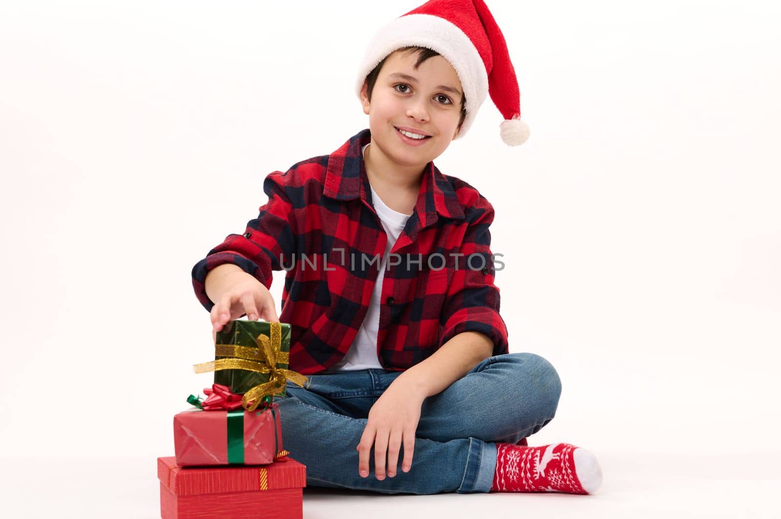 Happy Middle-Eastern preteen boy in Santa hat, red socks with Christmas pattern, smiles looking at camera while folding presents one on one, on a white background. Copy space. Happy Winter holidays