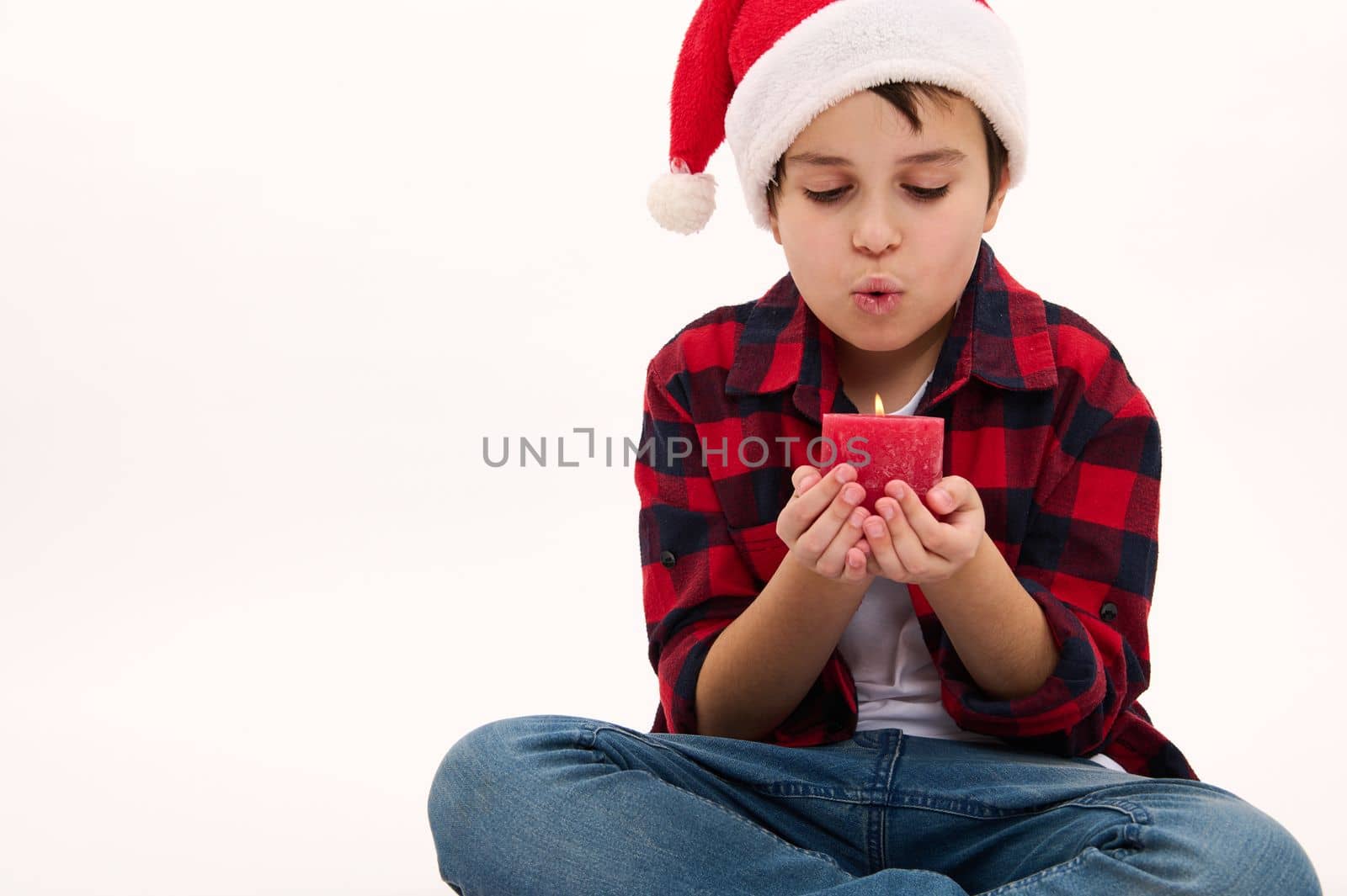 Lovely Caucasian child boy in Santa hat, makes a cherished wish for Christmas, blowing out a red candle, isolated on white background. Copy space. Enjoy magic mood of winter holidays. Happy childhood