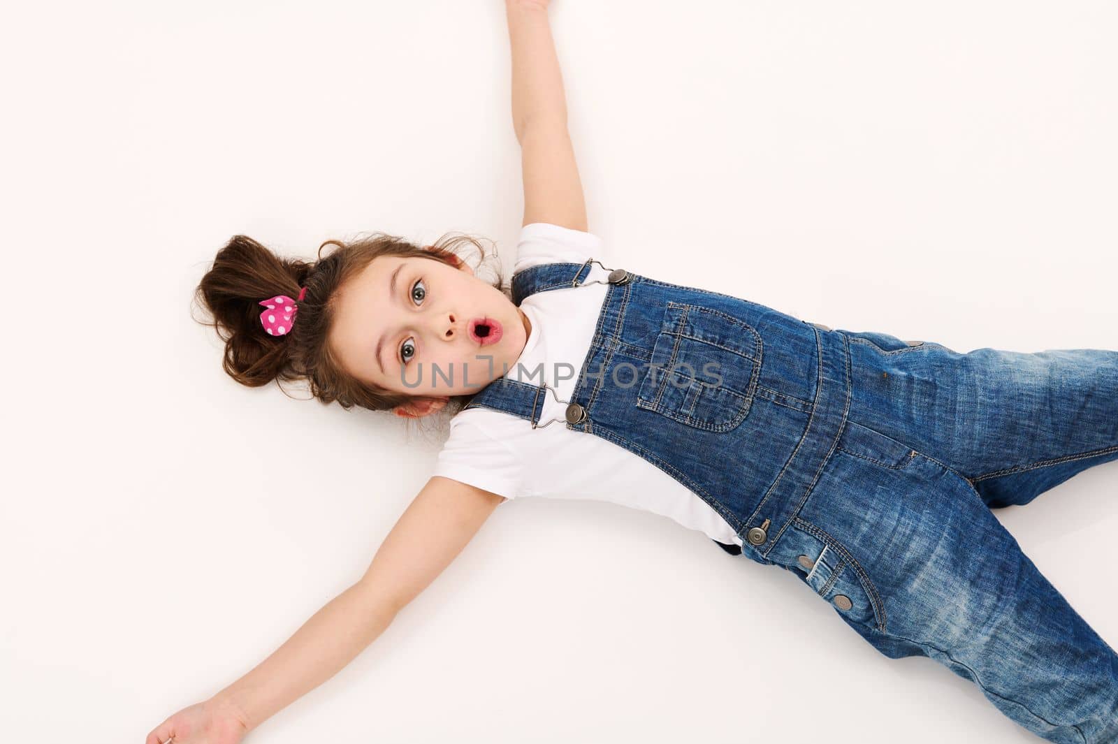 View from above of a happy little child girl, wearing blue denim overalls, experiencing astonishment surprise looking at camera, lying down on a white background with copy advertising space for text