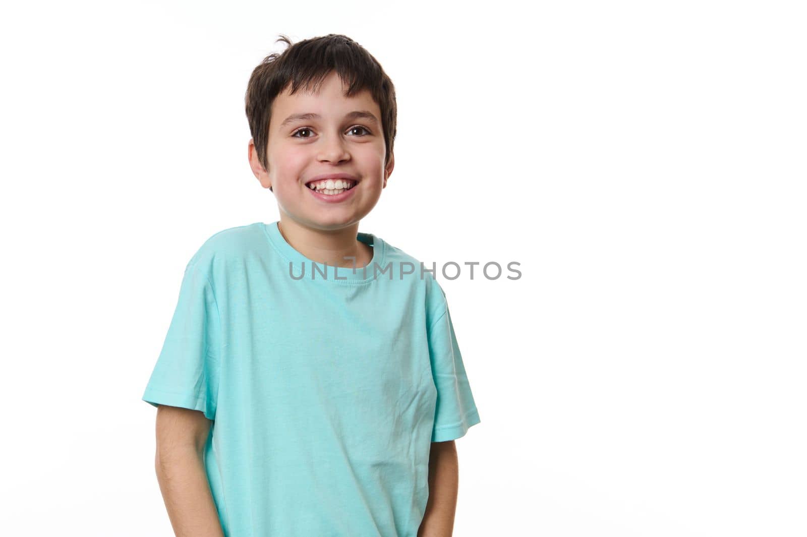 Emotional portrait of Caucasian cheerful teenage boy wearing turquoise t-shirt, smiling a beautiful toothy smile looking at camera, isolated on white background. Copy space for your promotional text