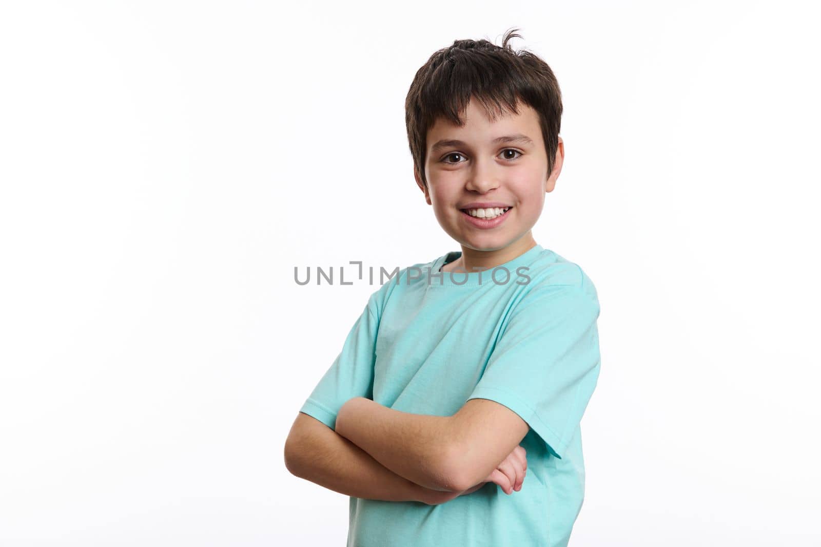 Handsome cheerful pre-adolescent boy smiling with beautiful toothy smile, confidently looking at camera, posing with crossed arms over white background with advertising space for you promotional text