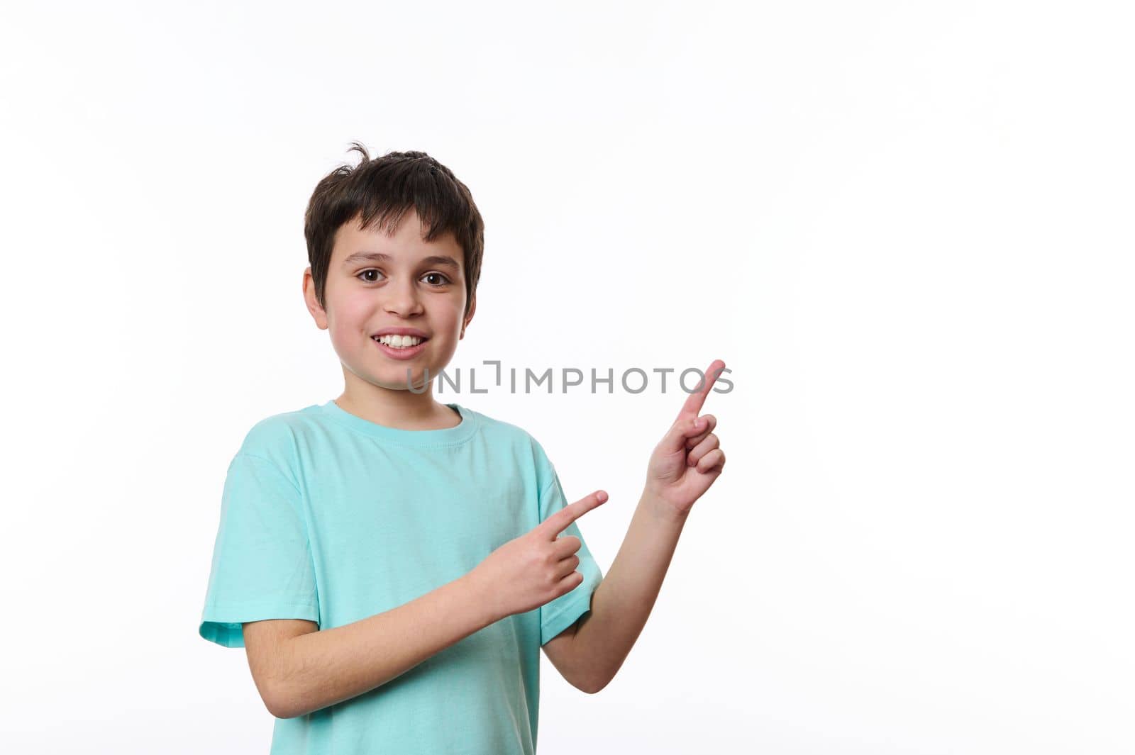 Portrait of an adorable smiling multi-ethnic preteen child, smart school age boy wearing blue turquoise t-shirt, looking at you while pointing at space for your advertising text on white background