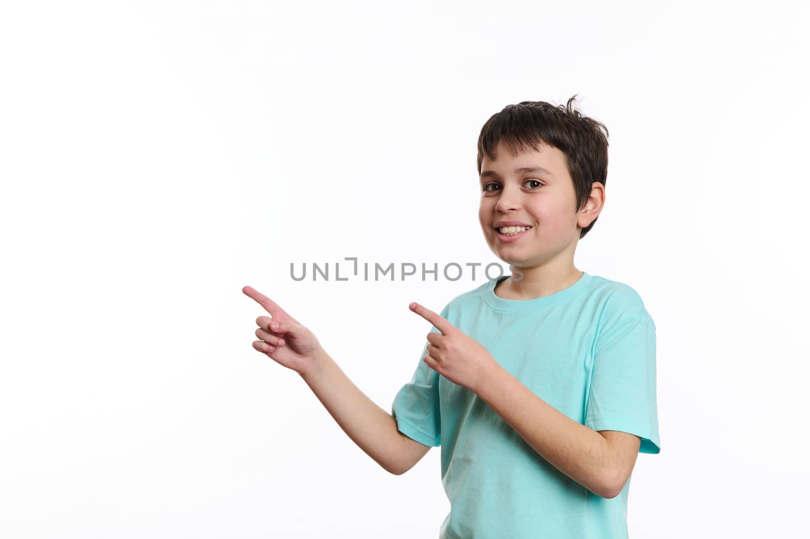 Isolated portrait on white background of a smart school boy, cheerfully smiling teenager wearing blue turquoise t-shirt, looking at you while pointing at advertising space for your promotional text