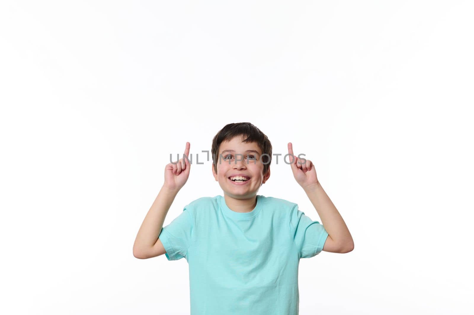 Portrait of Caucasian handsome pleasant pre-adolescent boy in blue casual t-shirt, smiling pleasantly, looking up, pointing finger at copy space for your advertising text, isolated on white background