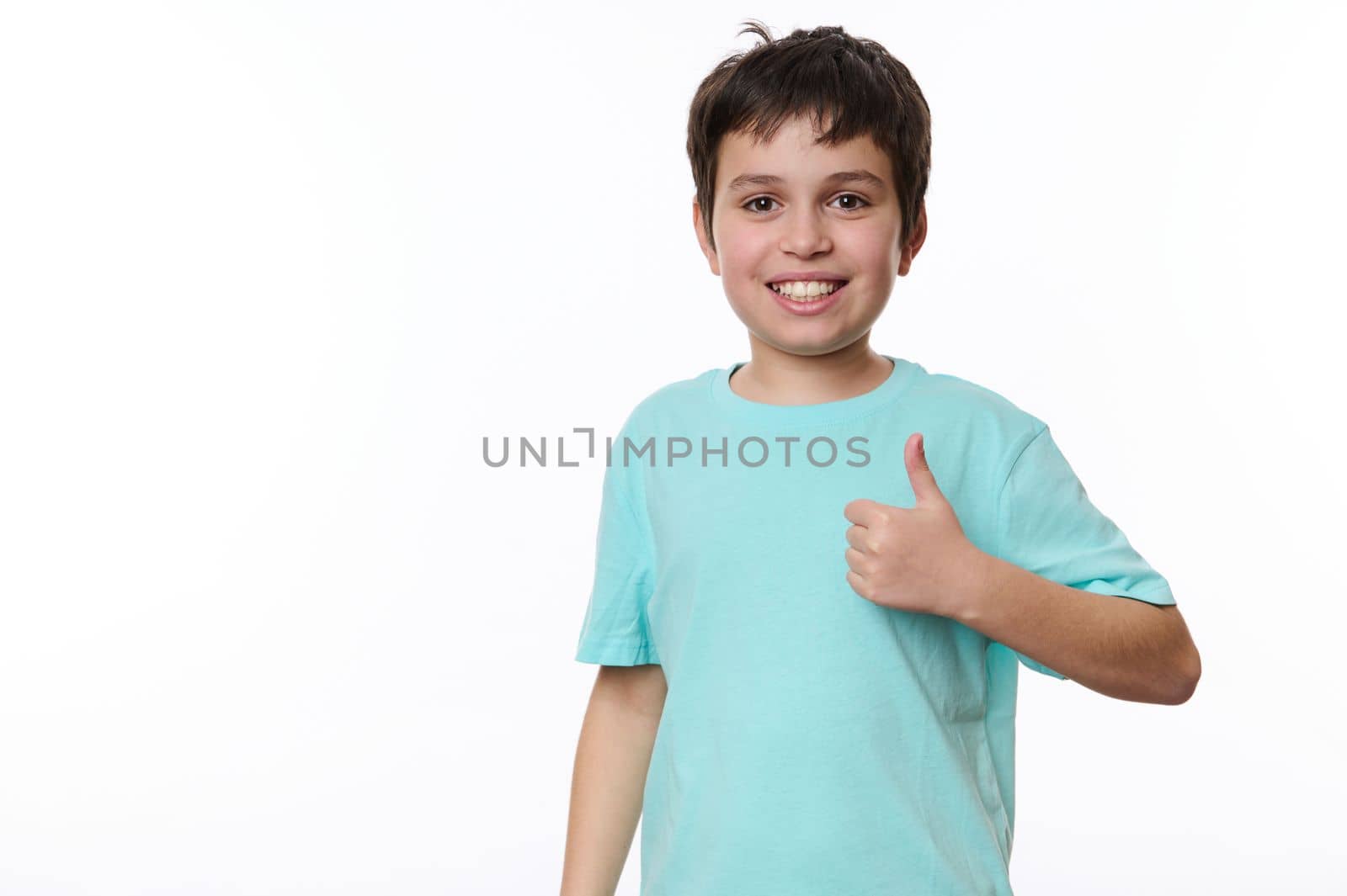 Happy Caucasian preteen child boy in blue t-shirt, a smart schoolboy shows a thumb up, cutely smiles looking at camera, isolated over white background. Free advertising space for your promotional text