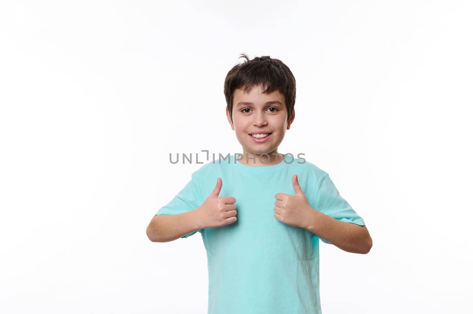Portrait of Caucasian handsome pleasant teenage child boy in blue casual t-shirt, smiling pleasantly, looking at camera, showing thumbs up, isolated on white background with space for promotional text