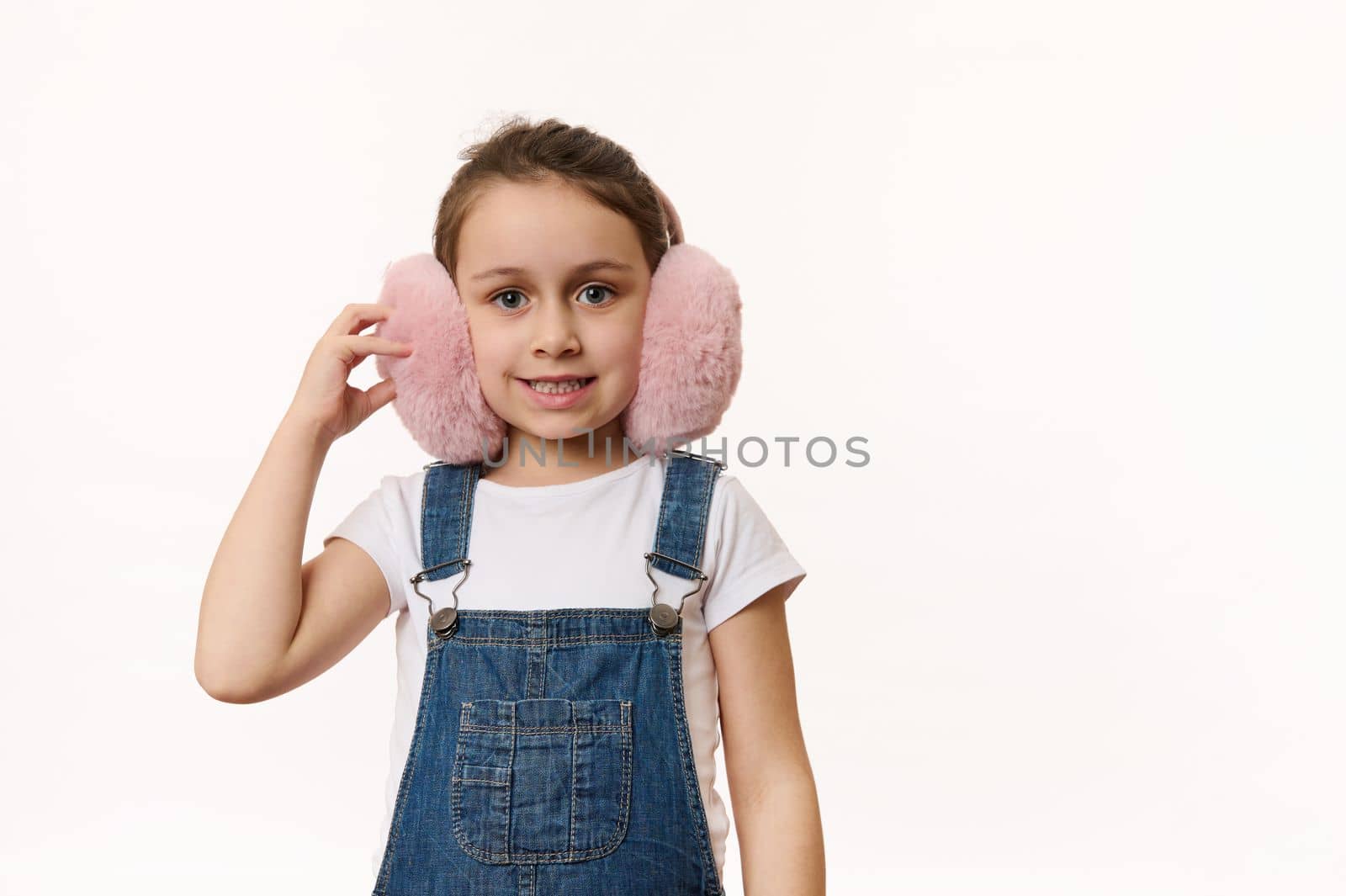 Happy child, lovely baby girl wearing cute pink winter earmuffs - plush headphones, cutely smiling looking at camera, isolated over white background. Protect your ears from cold. Copy space for text