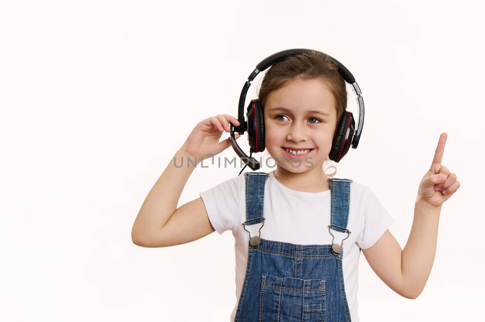Smart little school girl, first grader wearing audio headset, cutely smiles looking at camera, pointing at a copy space on white background. Online meeting, education. Distance learning. Video call.