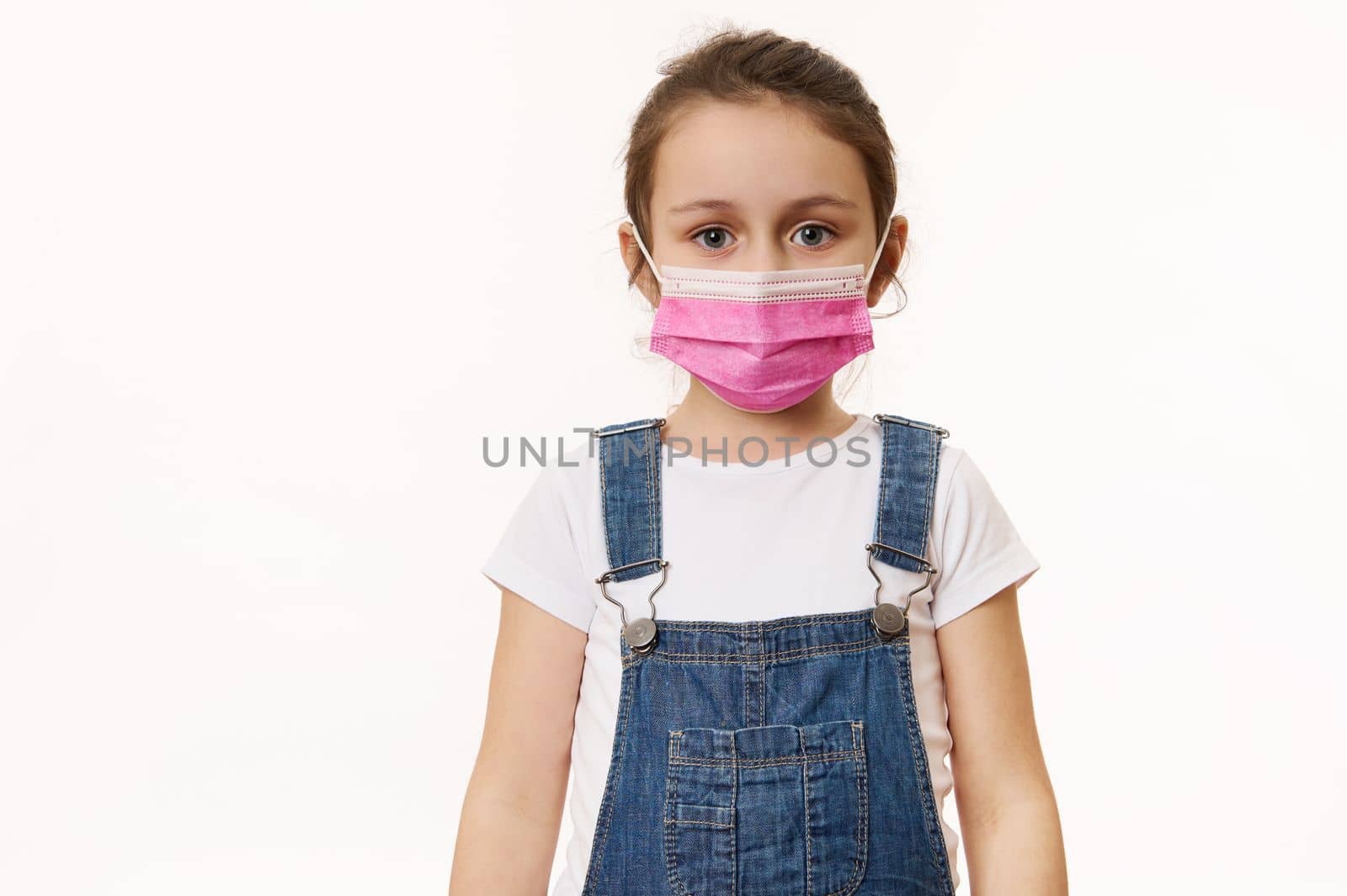 Beautiful European little girl in blue denim overalls, wearing a pink protective mask, isolated over white background. Free space for your advertising text. Pediatrics. Health care and medical content