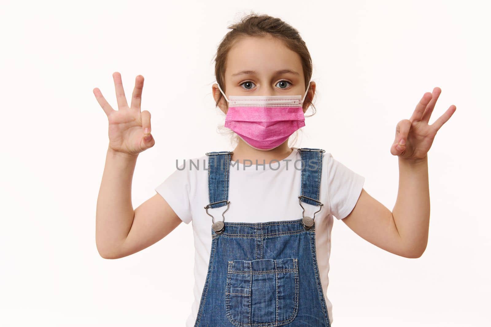 Adorable Caucasian little girl wearing pink protective medical mask, gestures with hands, shows OK signs, looks at camera, isolated over white background. Free space. Pediatrics. Health care. Medicine