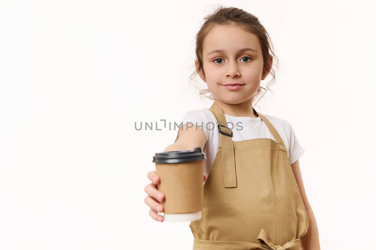 Caucasian lovely little girl wearing a beige chef's apron, handing out on camera a takeaway hot tea coffee drink in cardboard mug, isolated on white, copy space for text. Beverage in paper cup to go