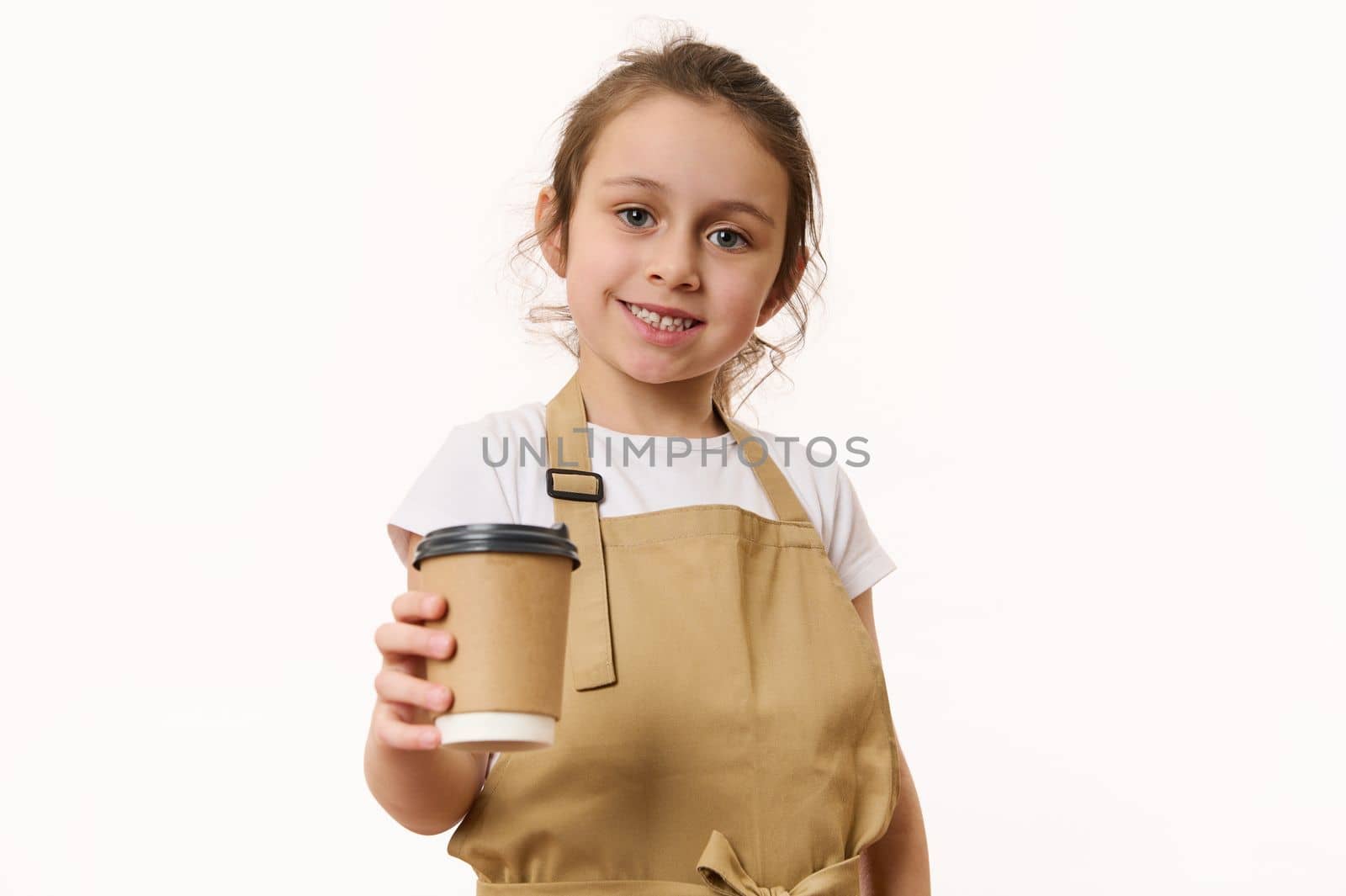Beautiful blue-eyed Caucasian little girl wearing a beige chef's apron, handing out a takeaway hot beverage, tea or coffee drink in an ecologically-friendly paper cup, cutely smiles looking at camera