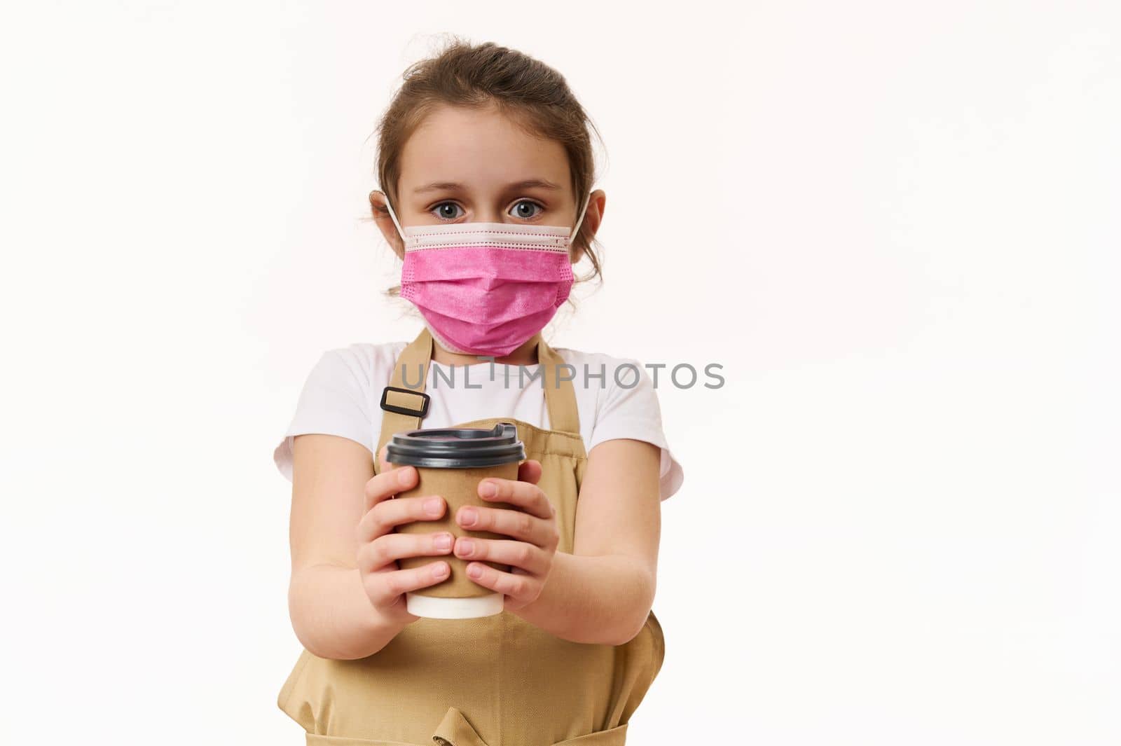 Caucasian little child girl wearing safety medical mask, dressed as barista, bartender with a hot drink in paper cup, isolated on white background. Cafe restaurant on pandemic lockdown. Takeaway food.