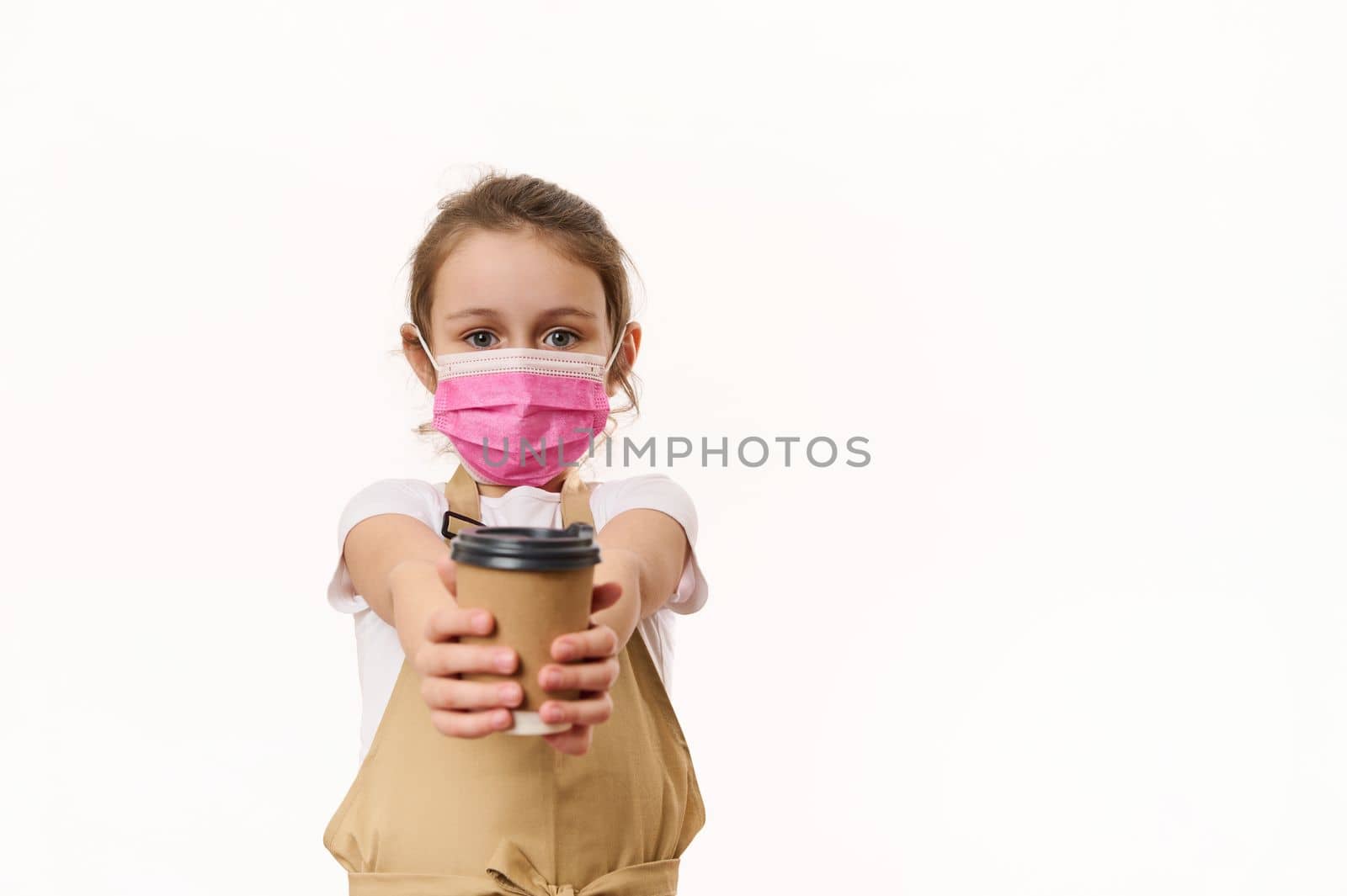 Caucasian lovely little girl wearing a beige chef's apron and pink medical protective mask, handing out on camera a takeaway hot tea cofee drink in paper mug, isolated on white, copy space for text