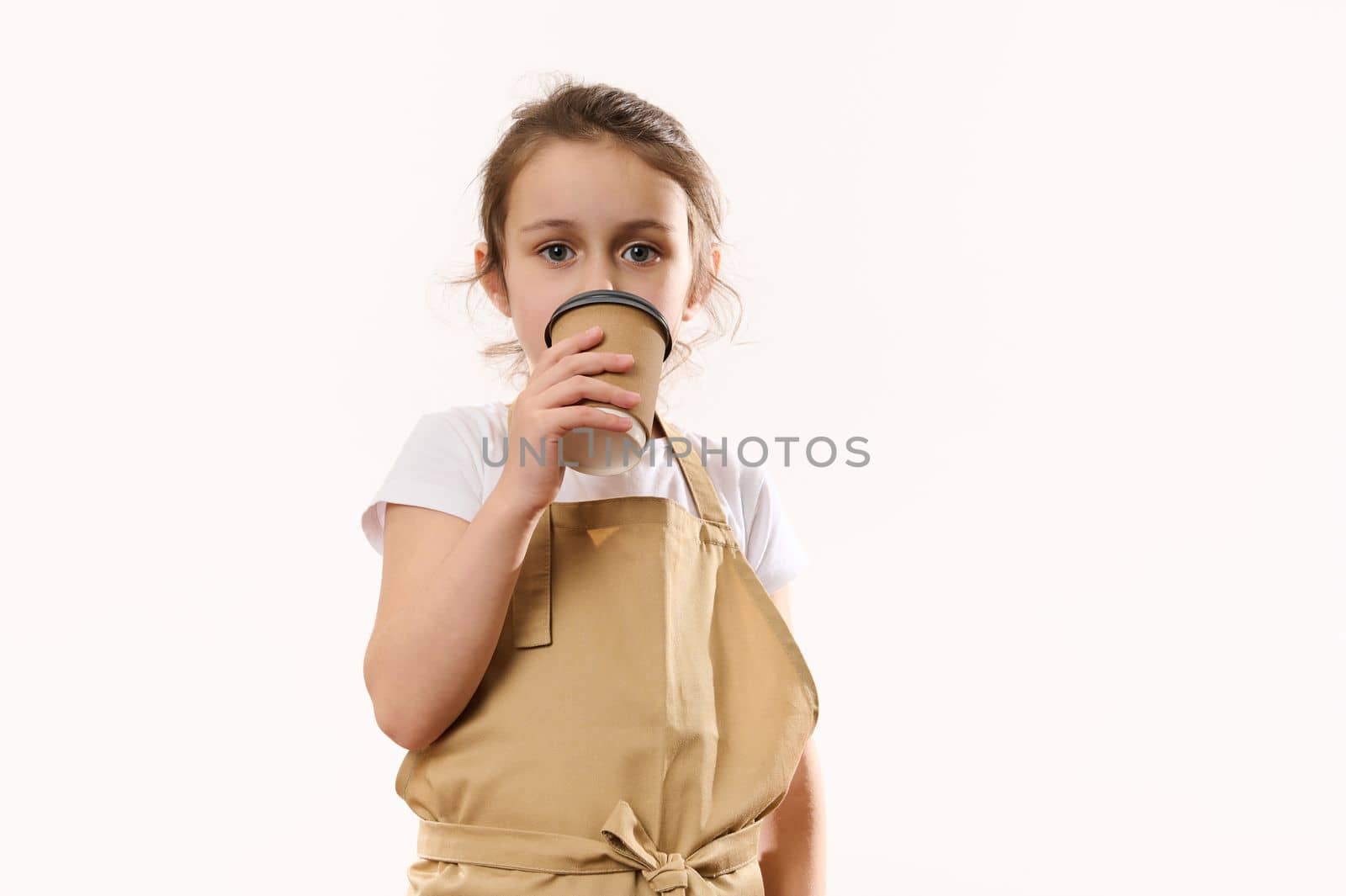 Caucasian beautiful little child girl dressed as barista bartender in beige chef's apron, drinks hot drink from an ecologically-friendly paper cup to go, smiling looking at camera, isolated on white