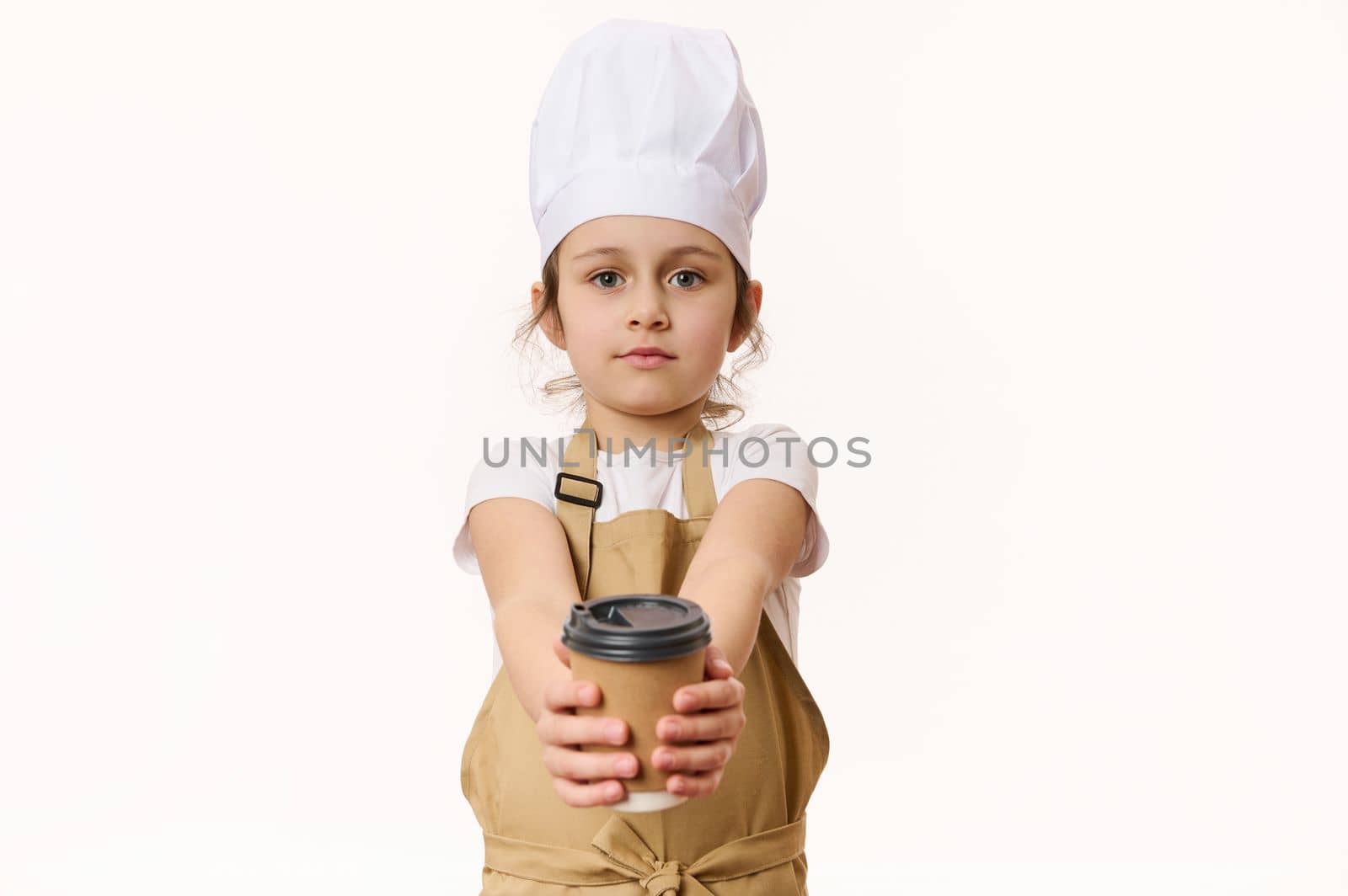Isolated portrait on white background of cute baby girl wearing a white chef's hat and beige apron, holding, selling a hot drink in a paper cup to go, looking at camera, isolated over white background