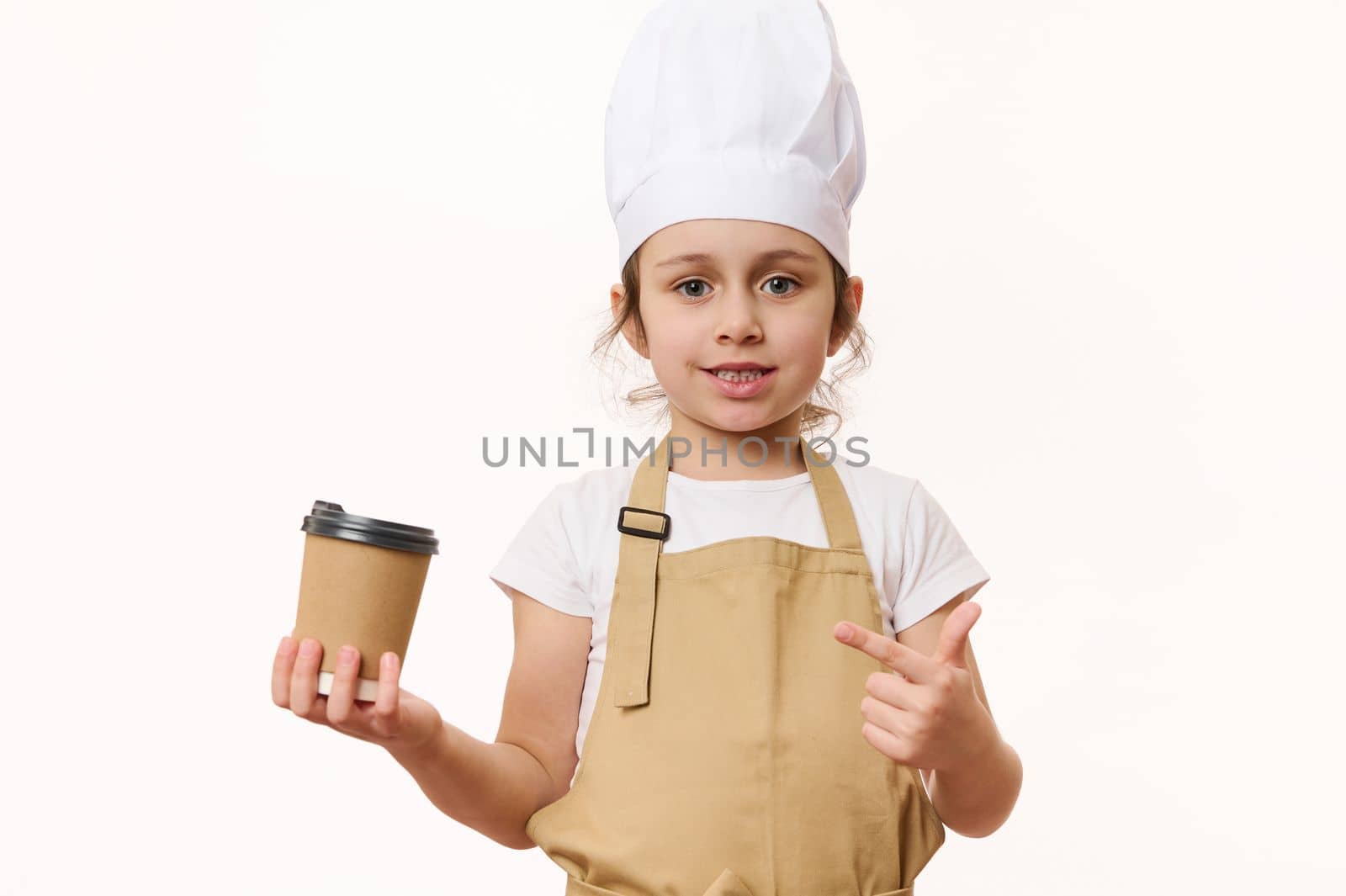 Cute baby girl - barista bartender in white chef hat and beige apron, smiling a beautiful toothy smile looking at camera, pointing at a hot drink in eco paper cup to go, isolated on white background