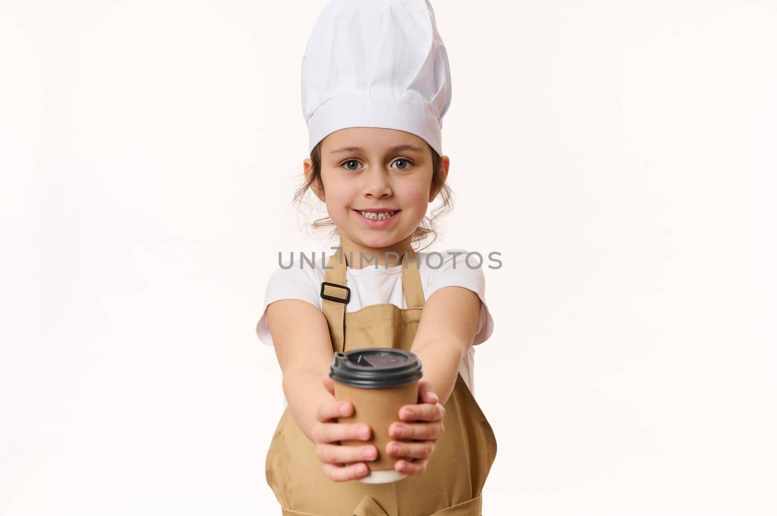 Caucasian little girl dressed as a barista, bartender in white chef hat and apron, smiling a beautiful toothy smile looking and handing on camera a hot drink in eco paper cup to go, isolated on white