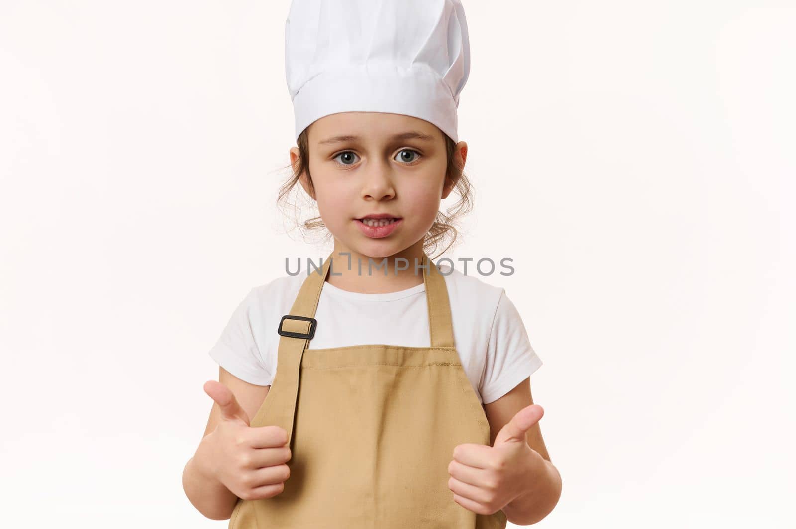 Isolated portrait on white background of adorable Caucasian child, lovely little girl - baker confectioner, cook wearing chef's hat and beige apron, shows thumbs up, looking at camera. Free ad space