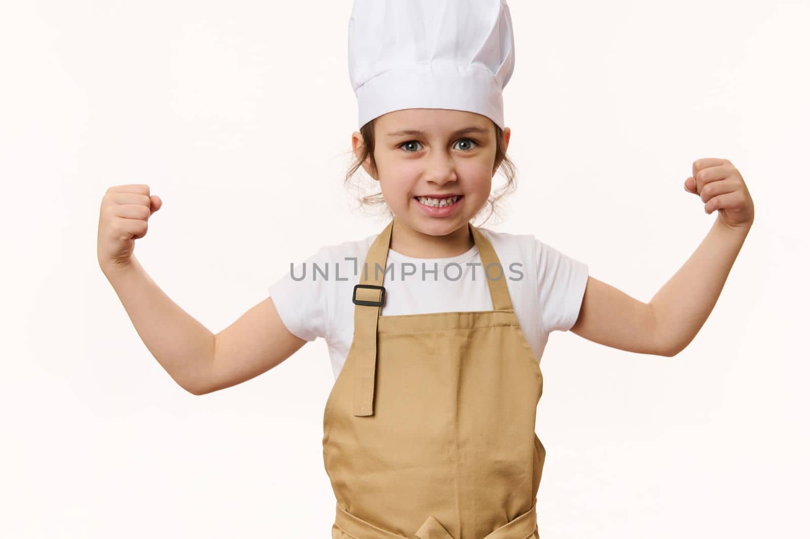 Delighted child, lovely baby girl dressed as pastry chef or baker confectioner, in hat and apron, clenching fists, looking at camera, isolated on white background with free space for advertising text
