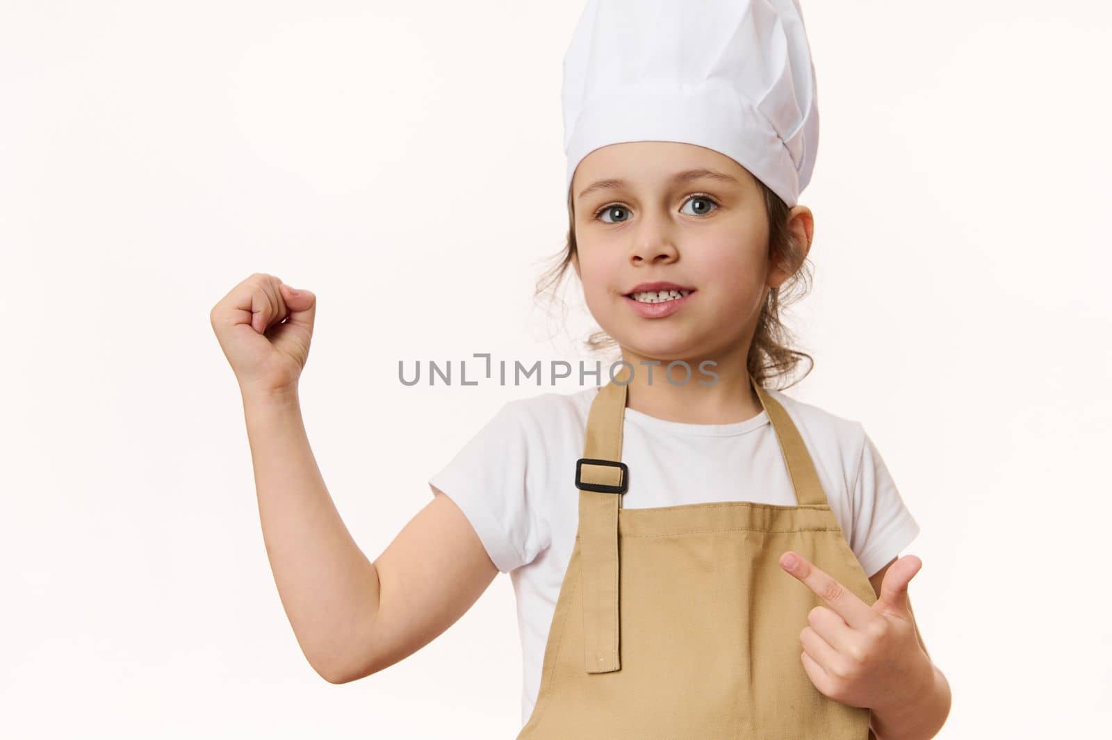 European beautiful little girl - chef pastry, cook, baker confectioner wearing kitchen apron and hat, isolated on white background with free space for promotional text. Cooking class for talented kids