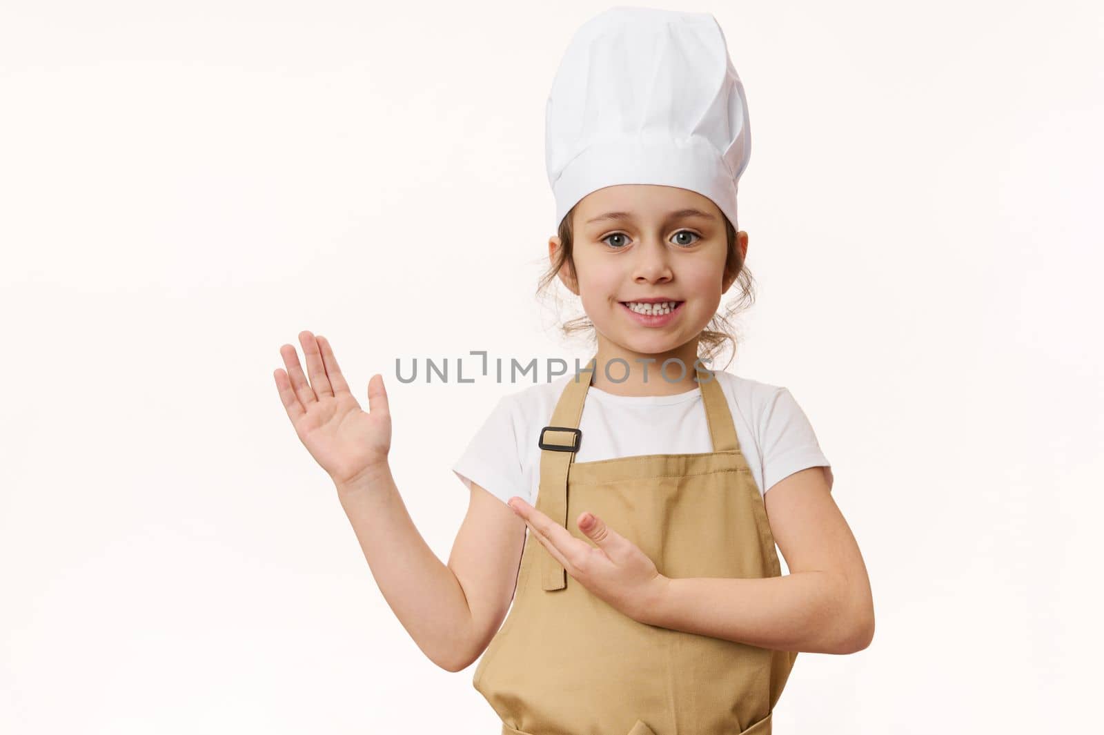 Charming little girl in chef's uniform, smiling sweetly at camera, pointing her hand at a blank place for advertising text, isolated on a white background. Concept for cooking class. Food consumerism