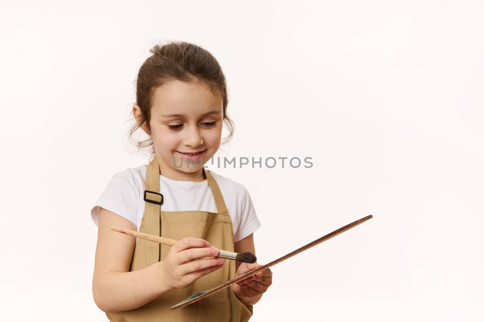 Inspired talented Caucasian little girl painter, artist wearing a beige apron, dipping a paintbrush on wooden palette with colorful oil paints, isolated on white background. Copy advertising space.