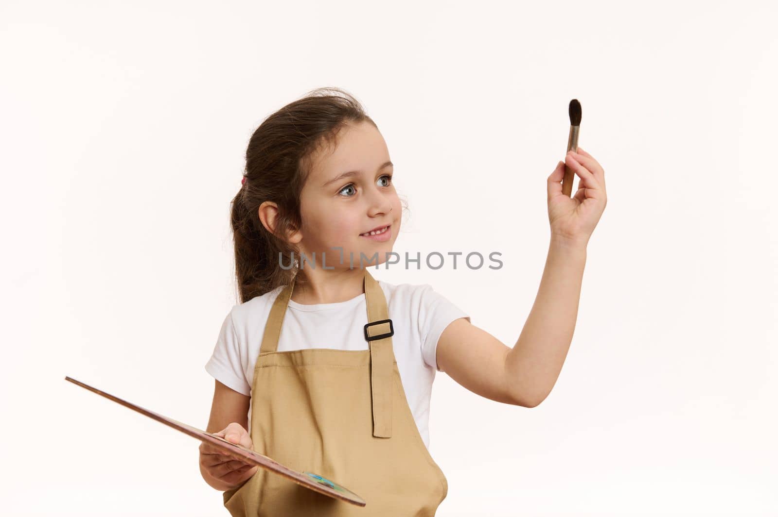Inspired little child artist, talented baby girl wearing beige apron, with a wooden palette of colorful oil paints and paintbrush, isolated over white background. Art. Creativity. Kids development