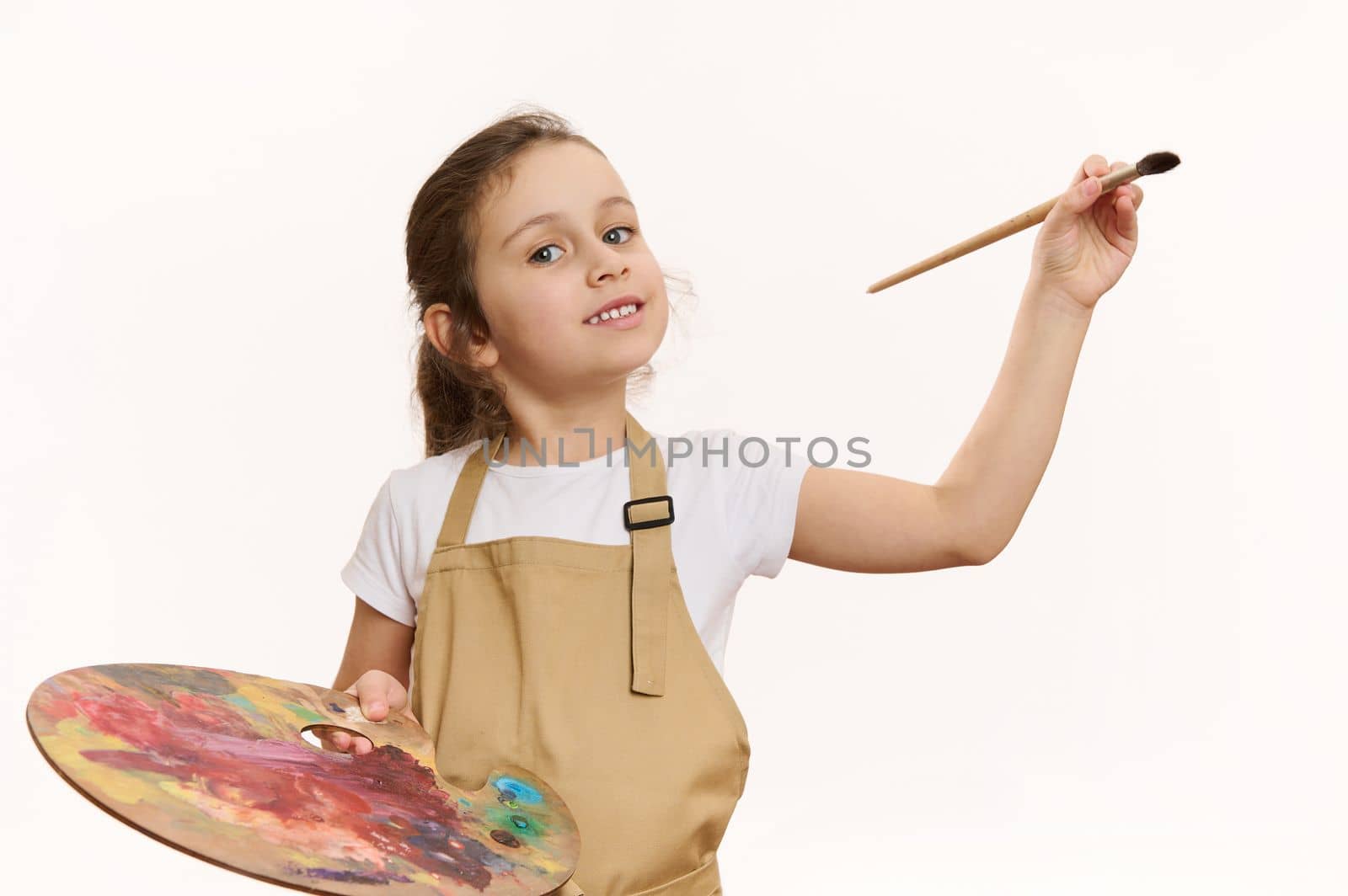 Lovely baby girl in beige apron, talented little painter with a paintbrush and palette of colorful paints, cutely smiling a toothy smile looking at camera, white background. Kids art development