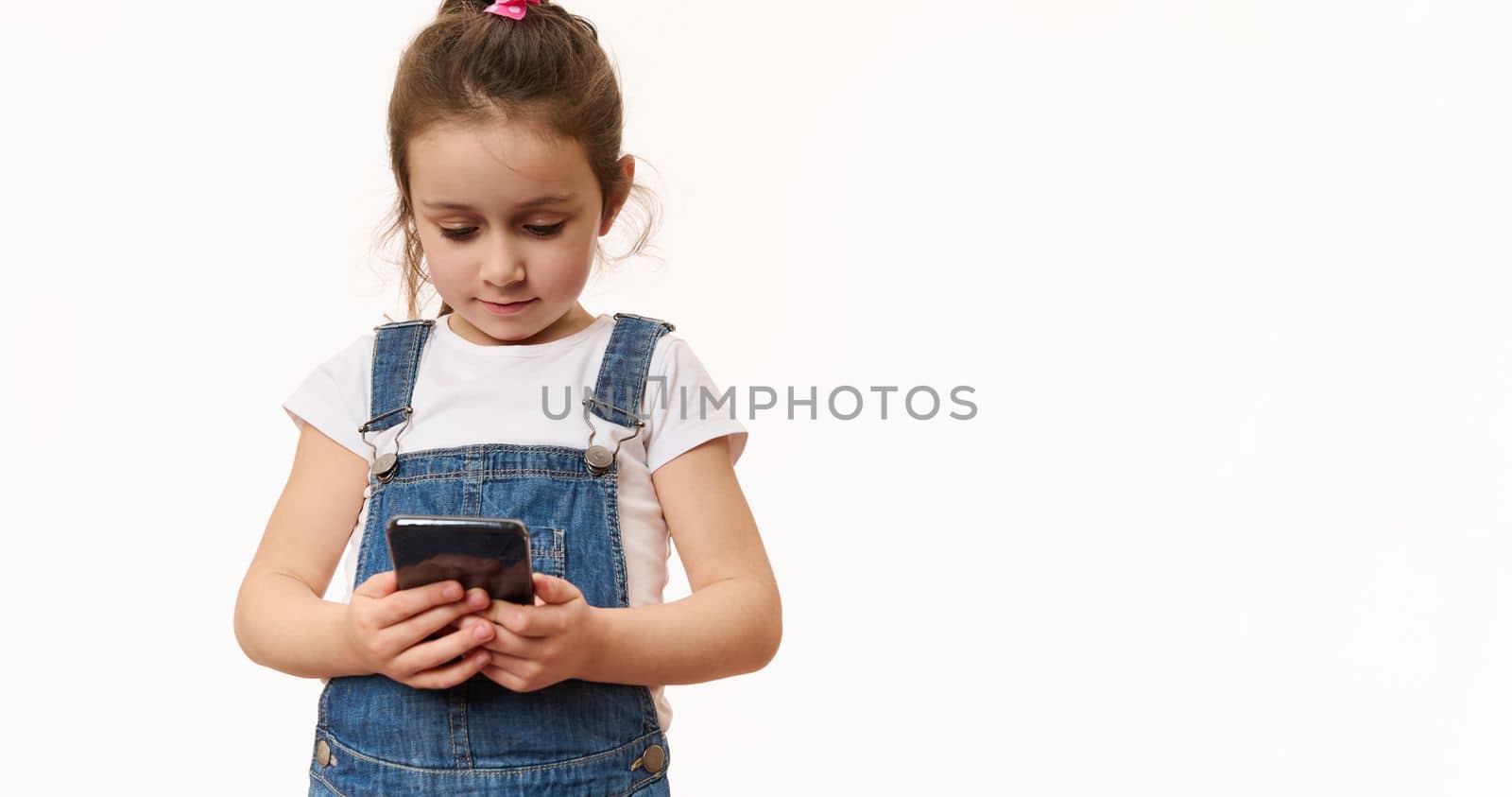 Isolated portrait on white background of a lovely little preschool girl, first grader with a smartphone. Adorable child using mobile phone, checks contents, makes call. Kids and digital technologies