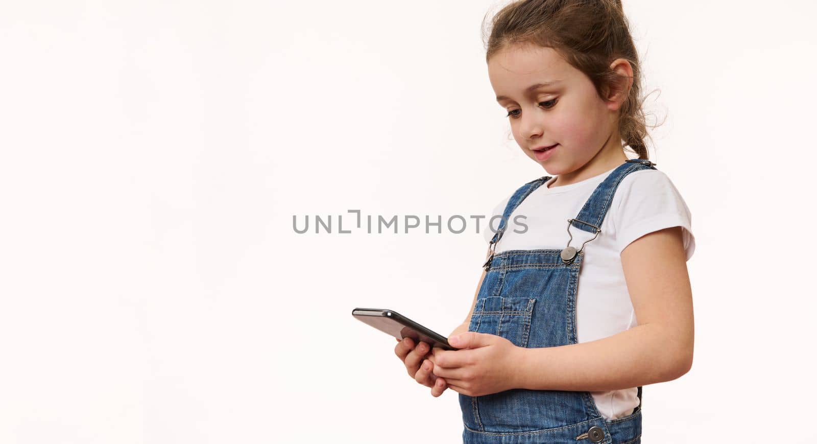 Studio shot on white background: Lovely child little girl using smartphone, plays game on mobile device, pleasantly surprised by easy application. Wow, great app for kids. Internet and digital gadgets