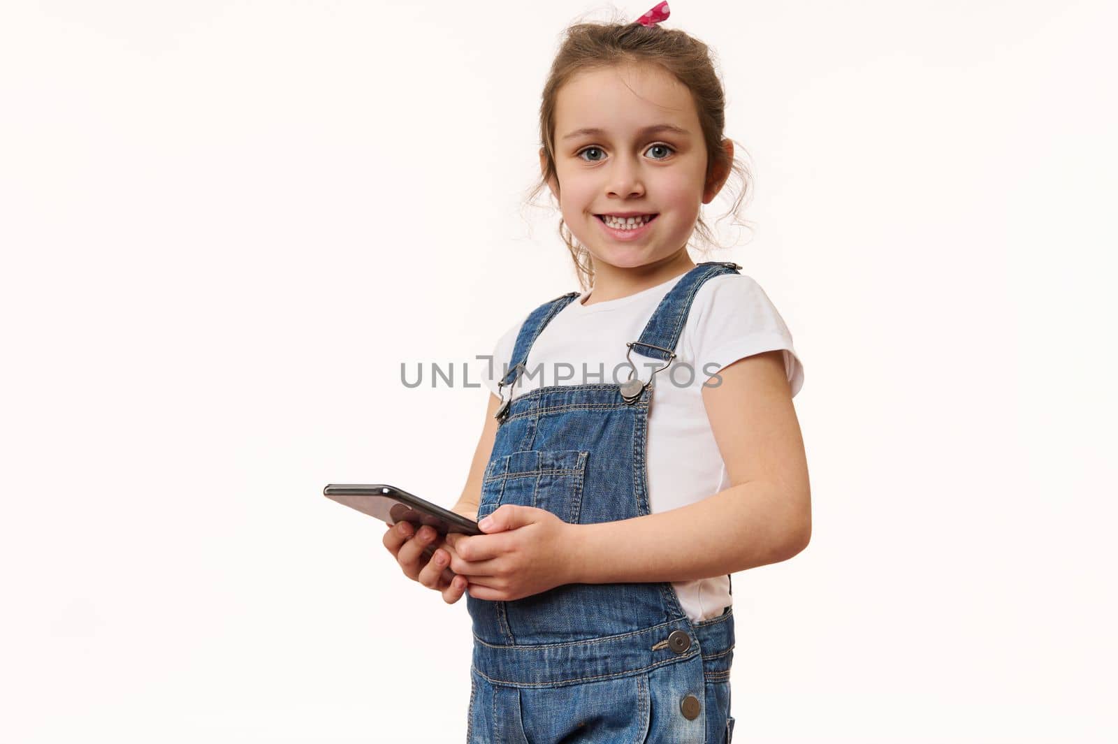 Adorable overjoyed little girl wearing blue denim overalls, holds a modern smartphone in her hands, cutely smiles looking at camera, posing isolated over white background with copy advertising space