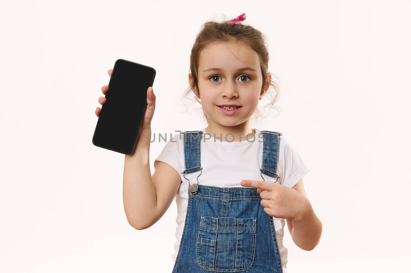 Wow awesome mobile app. Charming joyful cute little girl points at smartphone with black blank screen with copy space for young advertising text, smiles looking at camera, posing over white background