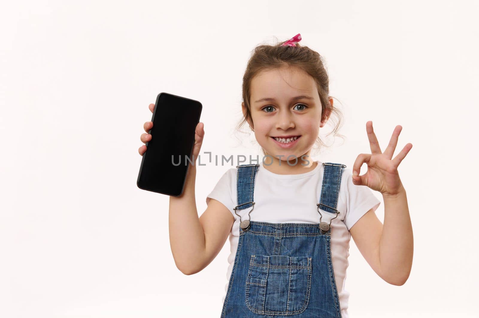 Lovely little kid, cute girl holding cell phone with blank screen with copy of advertising space for promo text, smiling with toothy smile, showing OK sign to camera. Children and digital gadgets