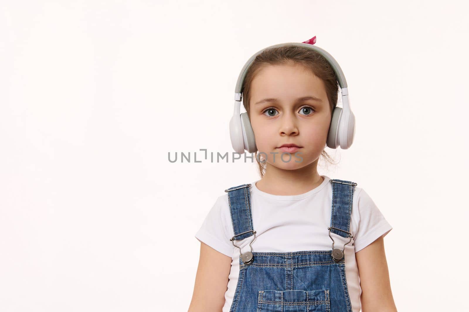 Adorable smart little child, cute baby girl wearing wireless headphones, looking at camera, isolated over white background. Studio shot with free space for your advertising and promotional text.