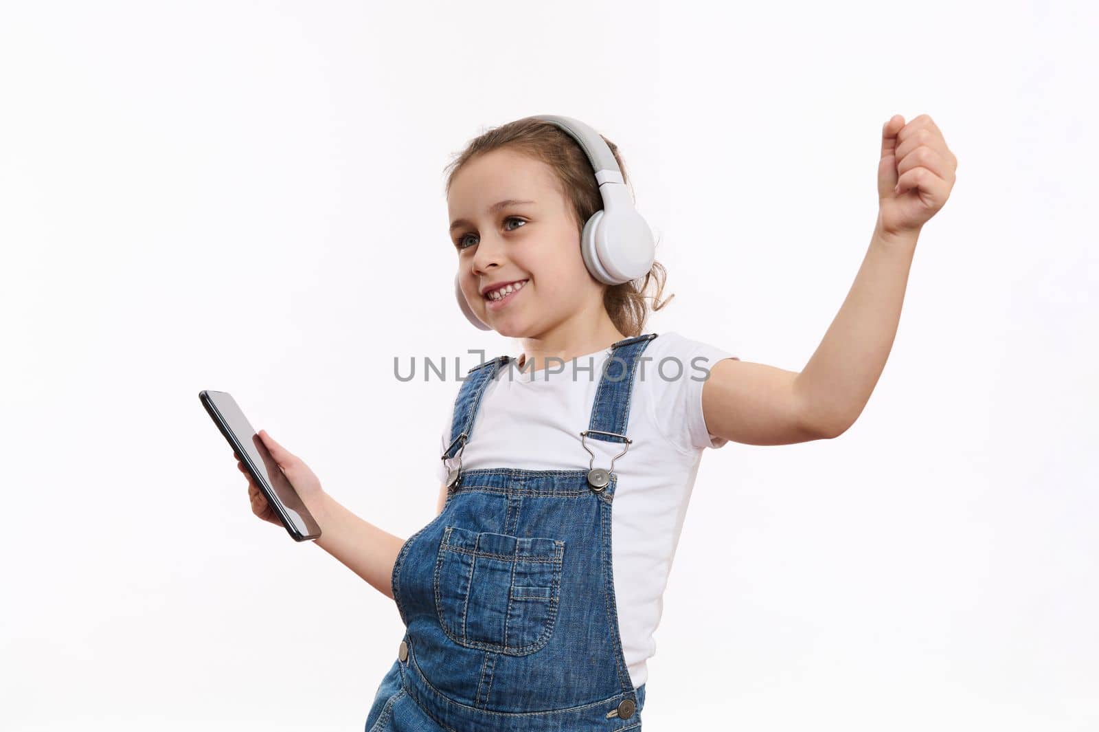 Joyful Caucasian child, a cute little girl wearing blue denim overalls, with smartphone in her hands, dances and listens to the music on her headphones, isolated over white background with copy space