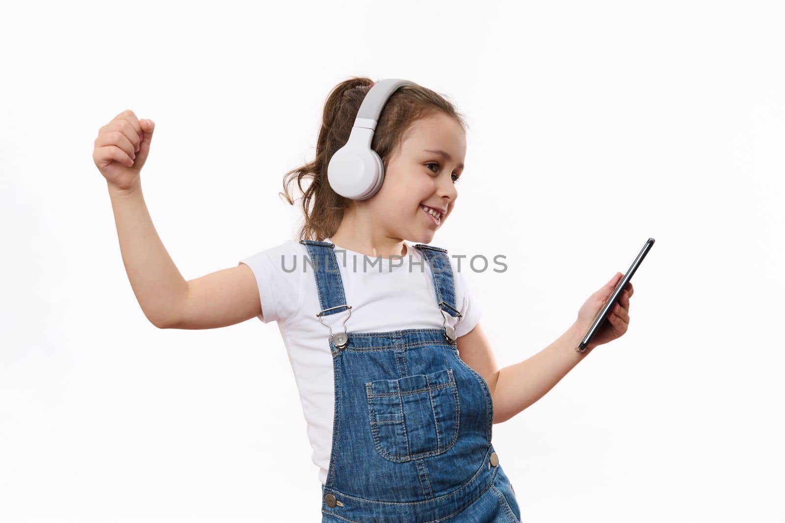 Adorable happy Caucasian little girl wearing blue denim overalls, holding mobile phone in her hands, dances and listens to the music on her stereo headphones, isolated white background, copy ad space
