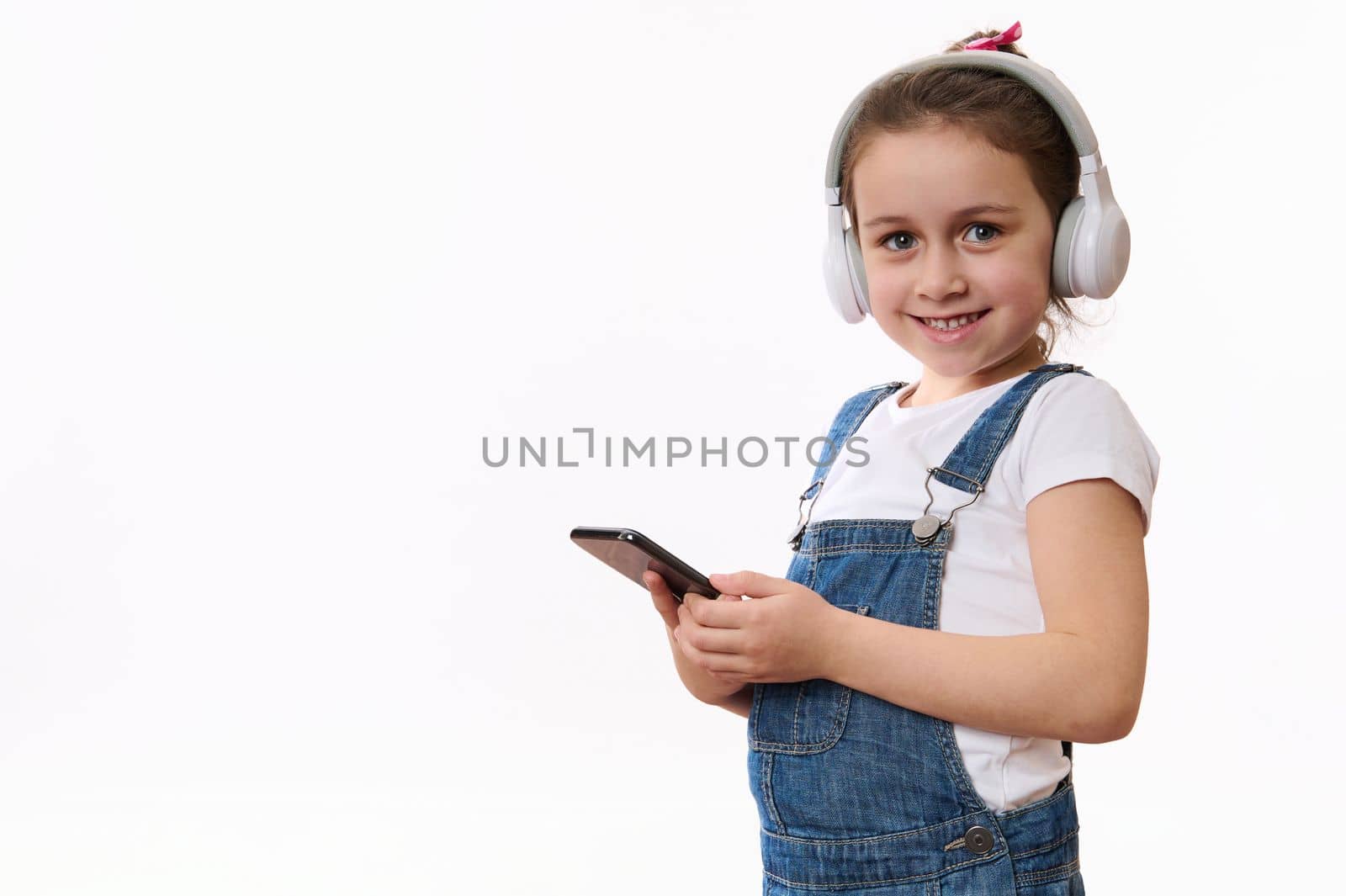Isolated portrait on white background of a Caucasian smart little child, adorable baby girl in denim overalls, wearing wireless headphones, holding mobile phone and cutely smiles looking at camera