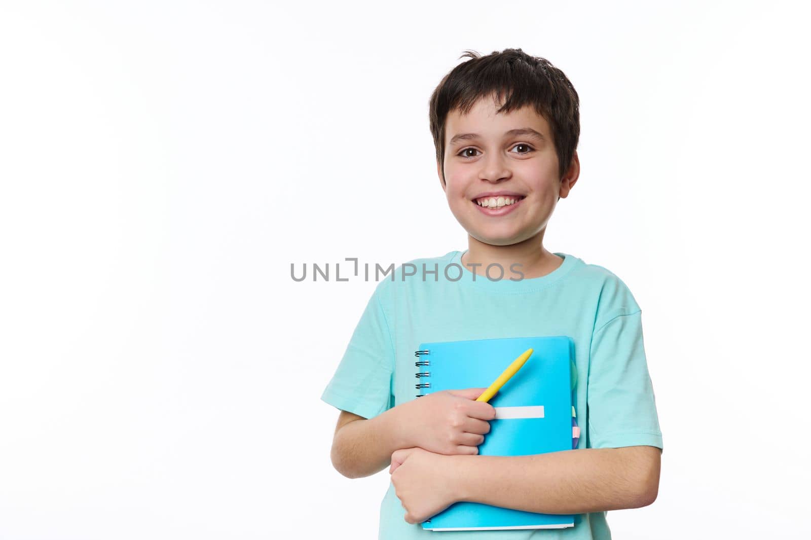 Lovely primary school student, smart teen boy in blue casual tshirt, holding textbook and yellow pen, smiling a cute toothy smile looking at camera, isolated, white background. Copy advertising space