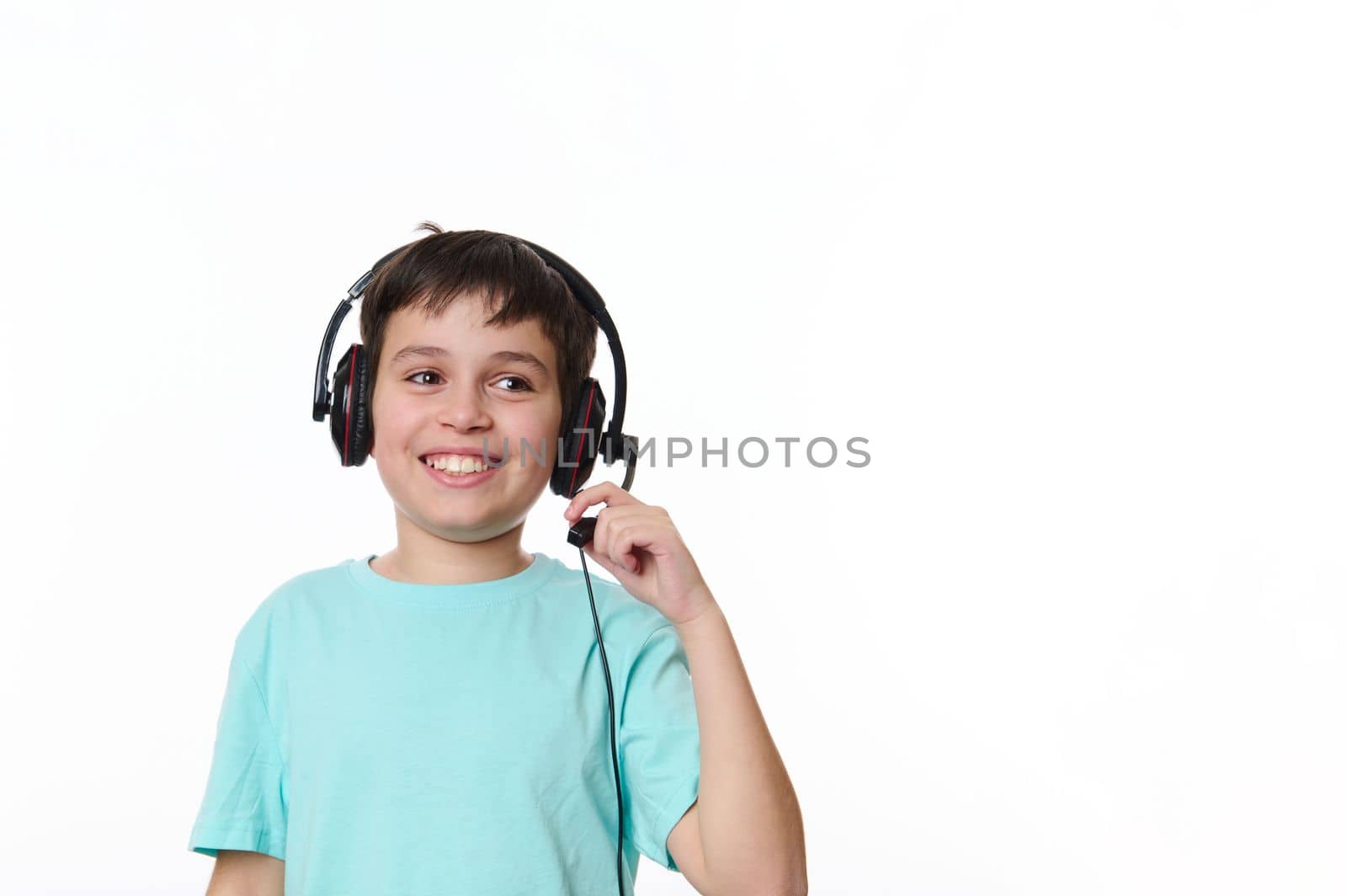 Adorable schoolboy wearing audio headset, headphones, smiling a cheerful toothy smile, looking aside at a copy space on white background. Online lesson via video link. child and technology concept