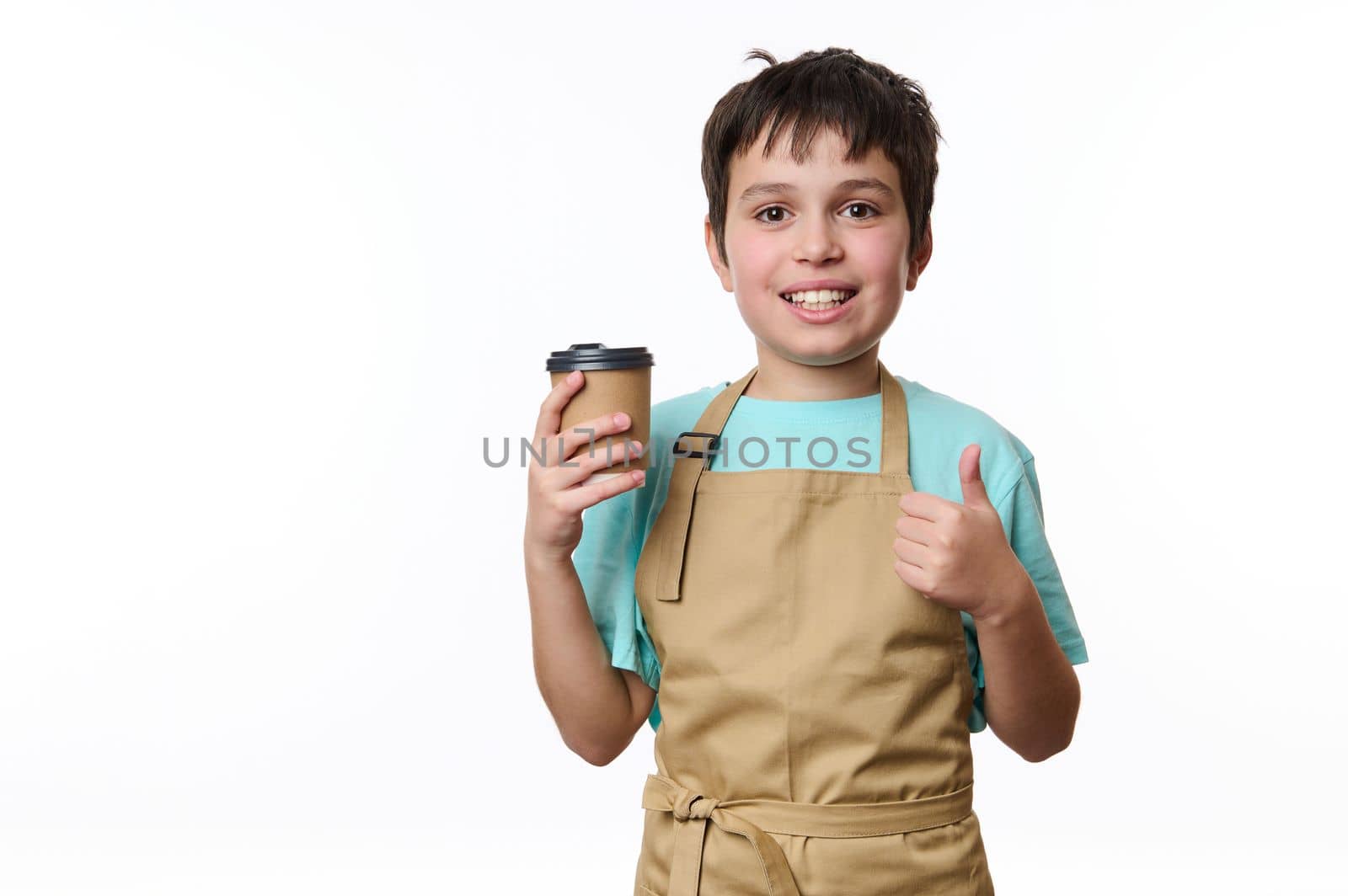 Isolated portrait on white background of a happy teenage boy bartender showing thumb up, selling a takeaway coffee beverage drink in cardboard mug. Hot beverage to go. Copy space for your advertising