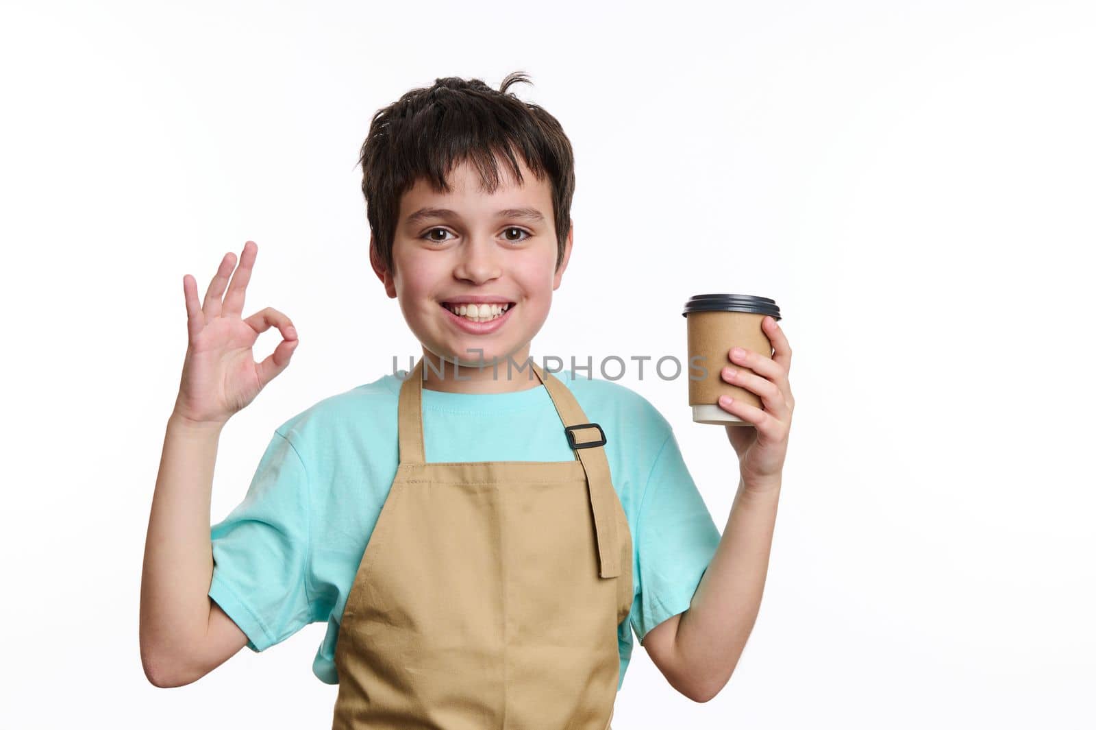 Amazing teenage boy - little barista bartender in chef apron, showing OK sign while selling hot takeaway drink in paper mug, smiling looking at camera, isolated on white background with free ad space