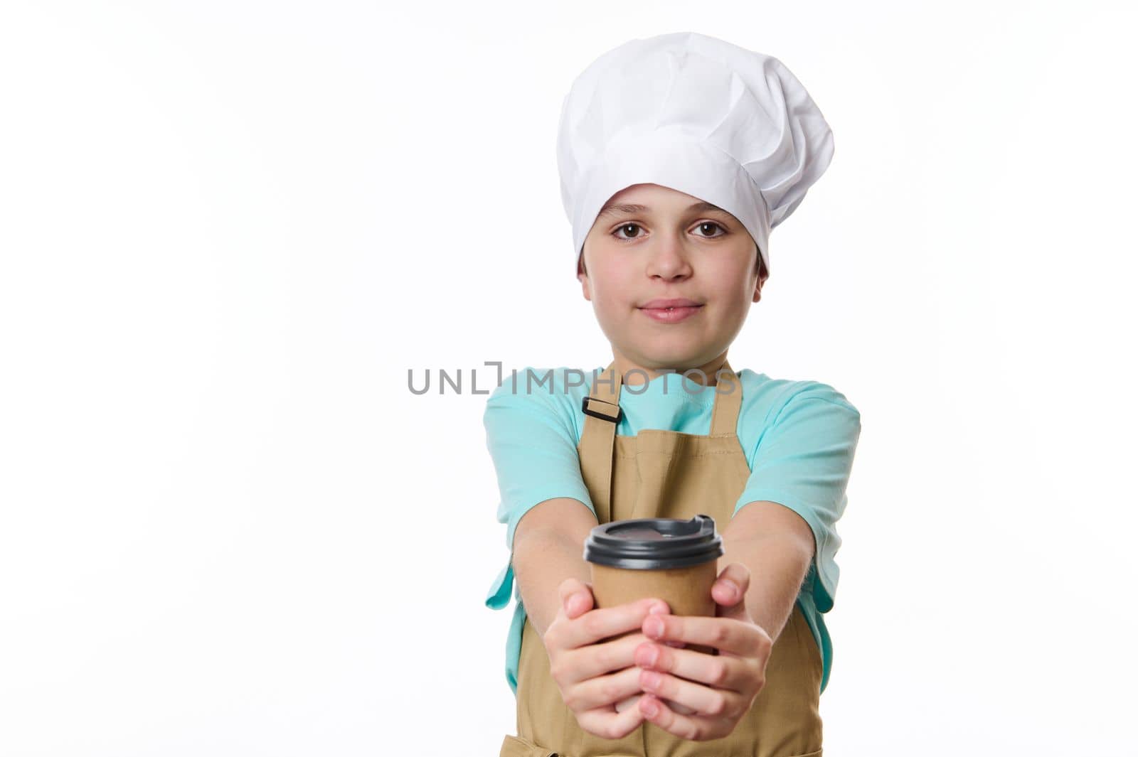 Multi-ethnic preteen boy bartender, wearing beige apron and white chef's hat, handing out on camera a takeaway hot tea coffee drink in cardboard mug. Beverage to go. Free space for promotional text.