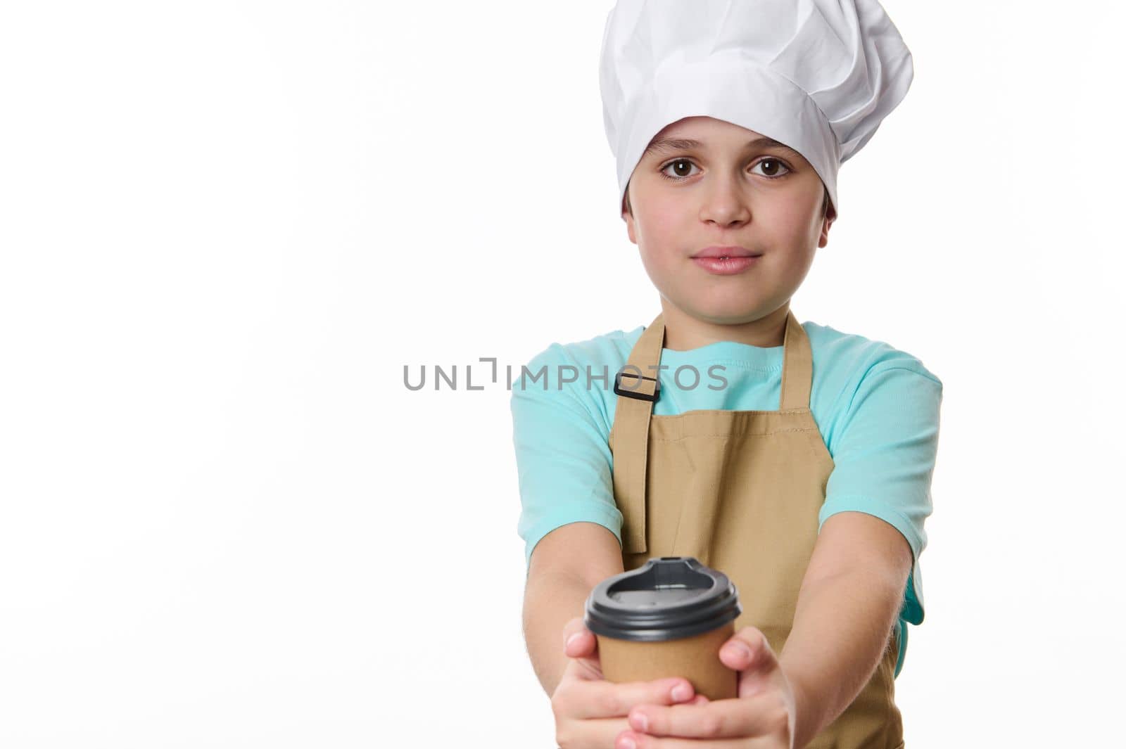 Cheerful teenage boy wearing a white chef's hat and beige apron, barista bartender handing hot coffee drink in a paper cup to go, isolated over white background. Free space for your advertising text