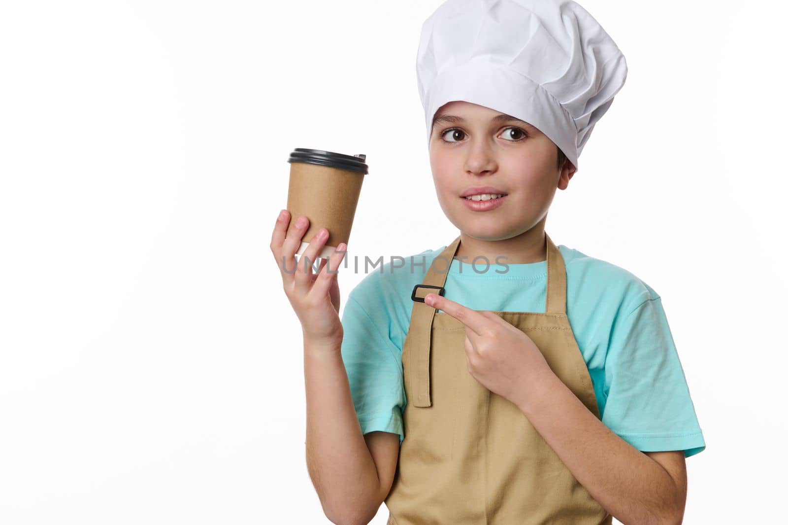 Hispanic handsome preteen boy dressed as bartender barista in beige chef apron and hat, points at a takeaway hot coffee tea drink in eco paper cup, isolated on white background. Free advertising space