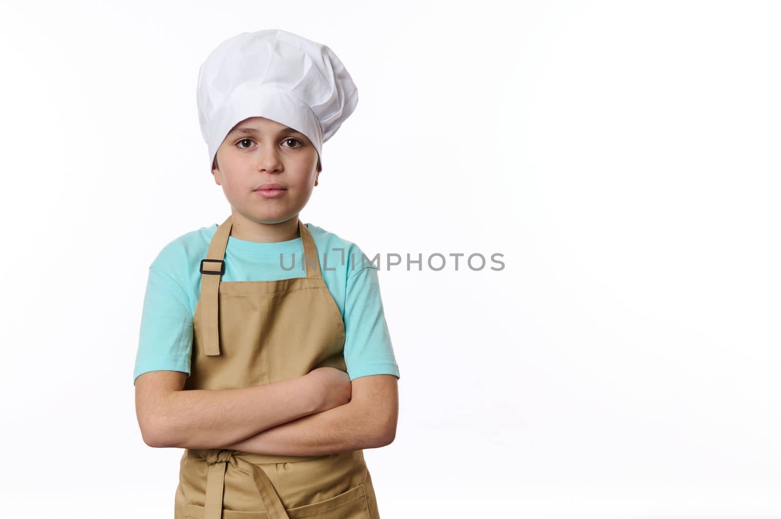 Confident portrait of a handsome teenage boy, dressed as a cook or baker confectioner in chef's hat and beige apron, isolated on white background with free space for your advertising text.
