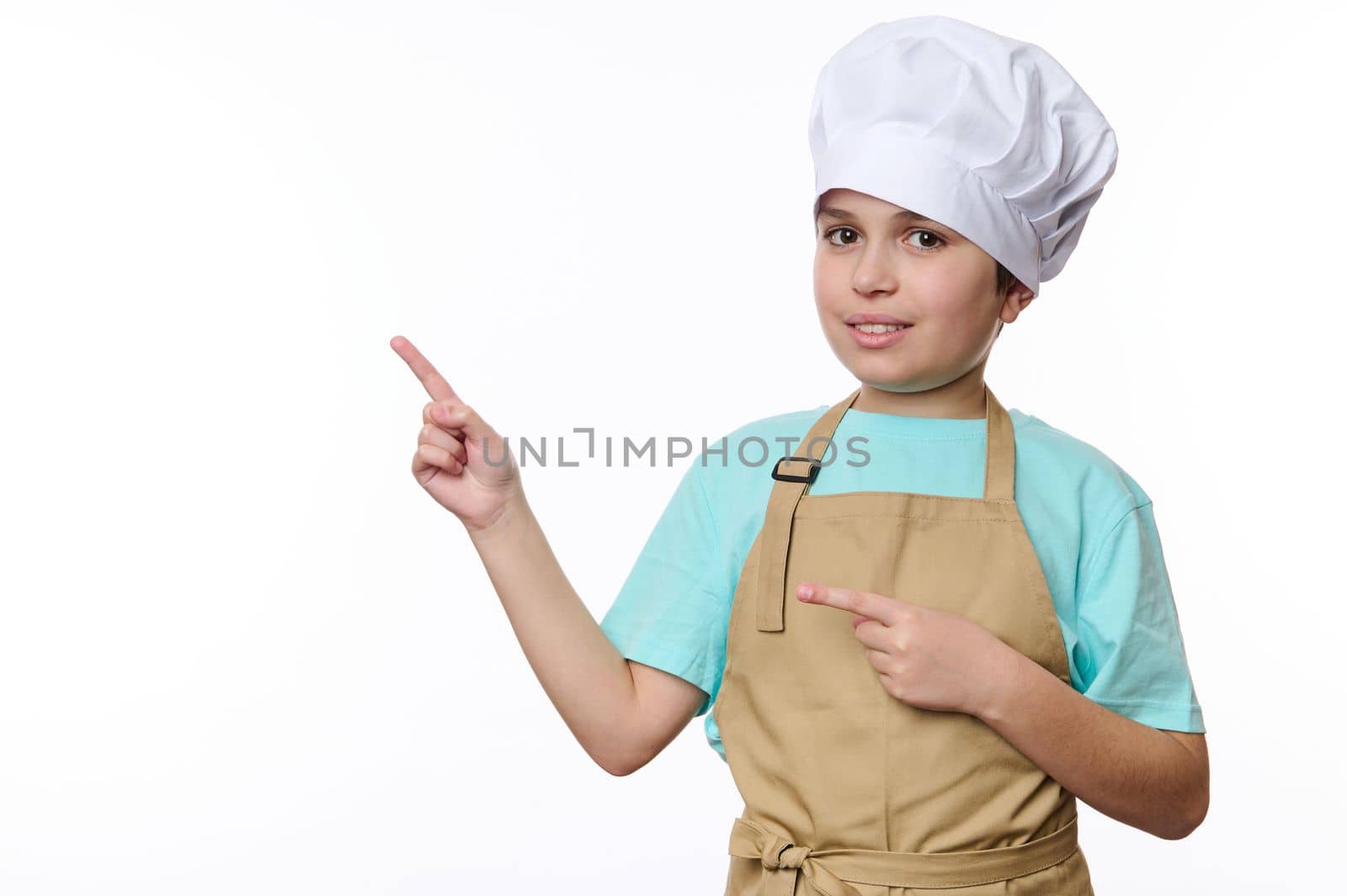 Adorable teenage boy dressed as a chef pastry or baker confectioner, wearing a white hat and beige apron, looks at camera and points at free space for promotional text, isolated over white background