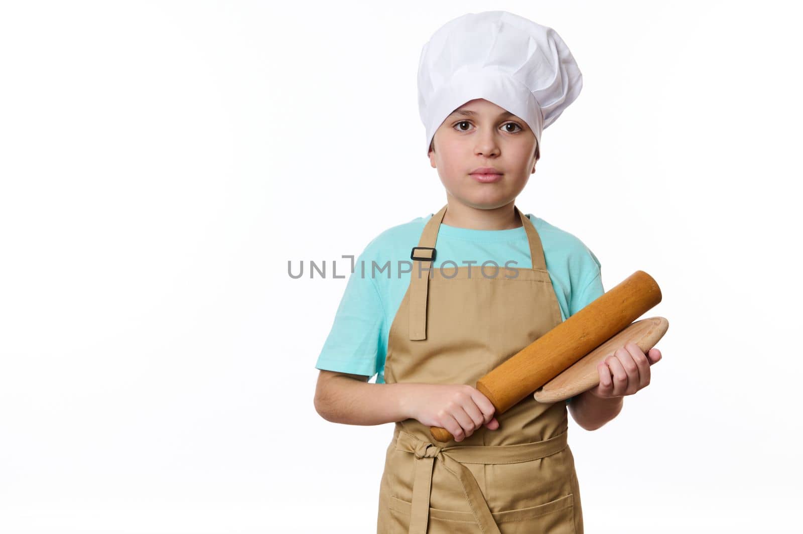 Isolated portrait on white background of a handsome teenage boy, wearing a white chef's hat and beige apron, a cook, baker confectioner posing with wooden board and rolling pin. Free advertising space