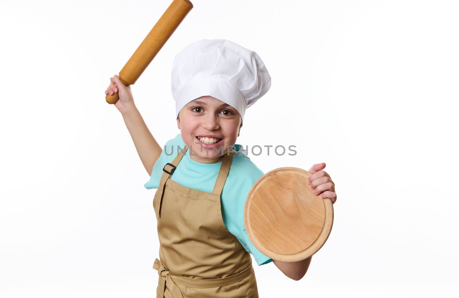 Isolated portrait on white background of a playful teenage boy, baker wearing chef's hat and apron, fighting with a wooden board and rolling pin in his hands, looking at camera. Free advertising space