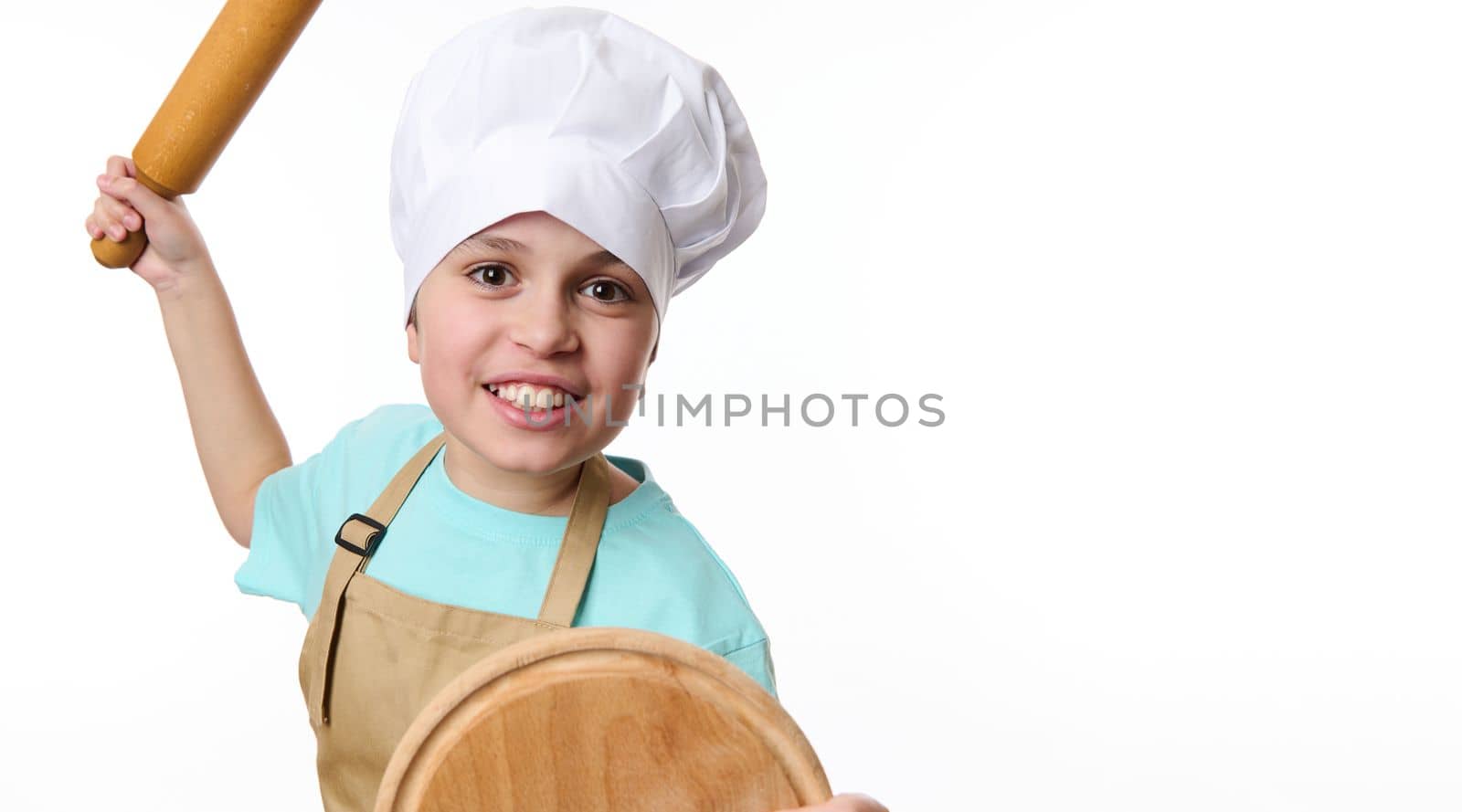 Close-up of a smiling mischievous boy - baker confectioner wearing chef's hat and apron, fighting with wooden board and rolling pin in his hands, isolated on white background. Bakery advertising shot.